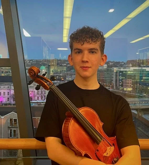 A young man with curly hair holding a viola in front of a large window with a cityscape view.