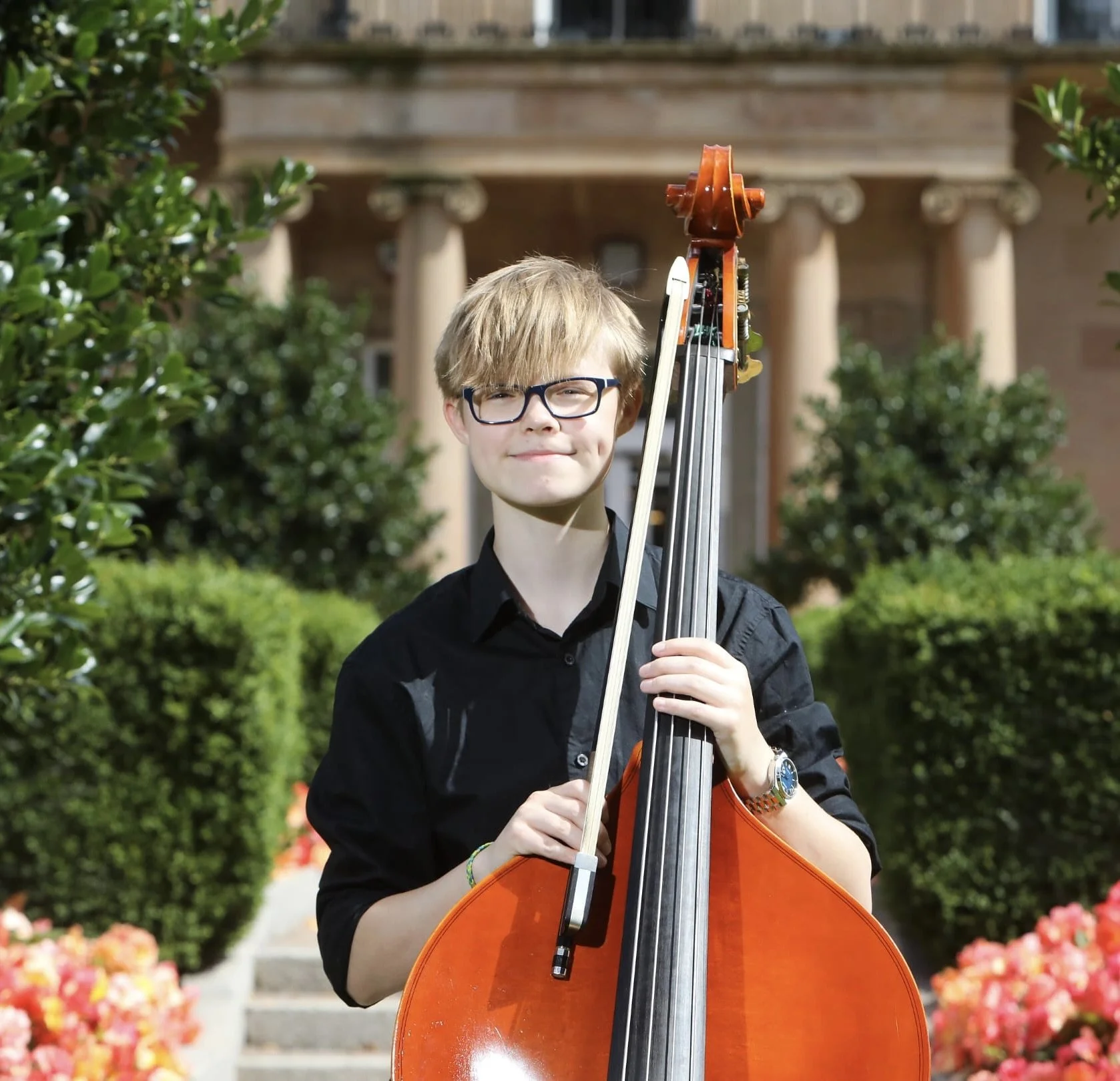 A young musician with blonde hair, glasses, and a black button-up shirt standing outdoors while holding a double bass, with greenery, flowers, and steps leading to a building in the background.