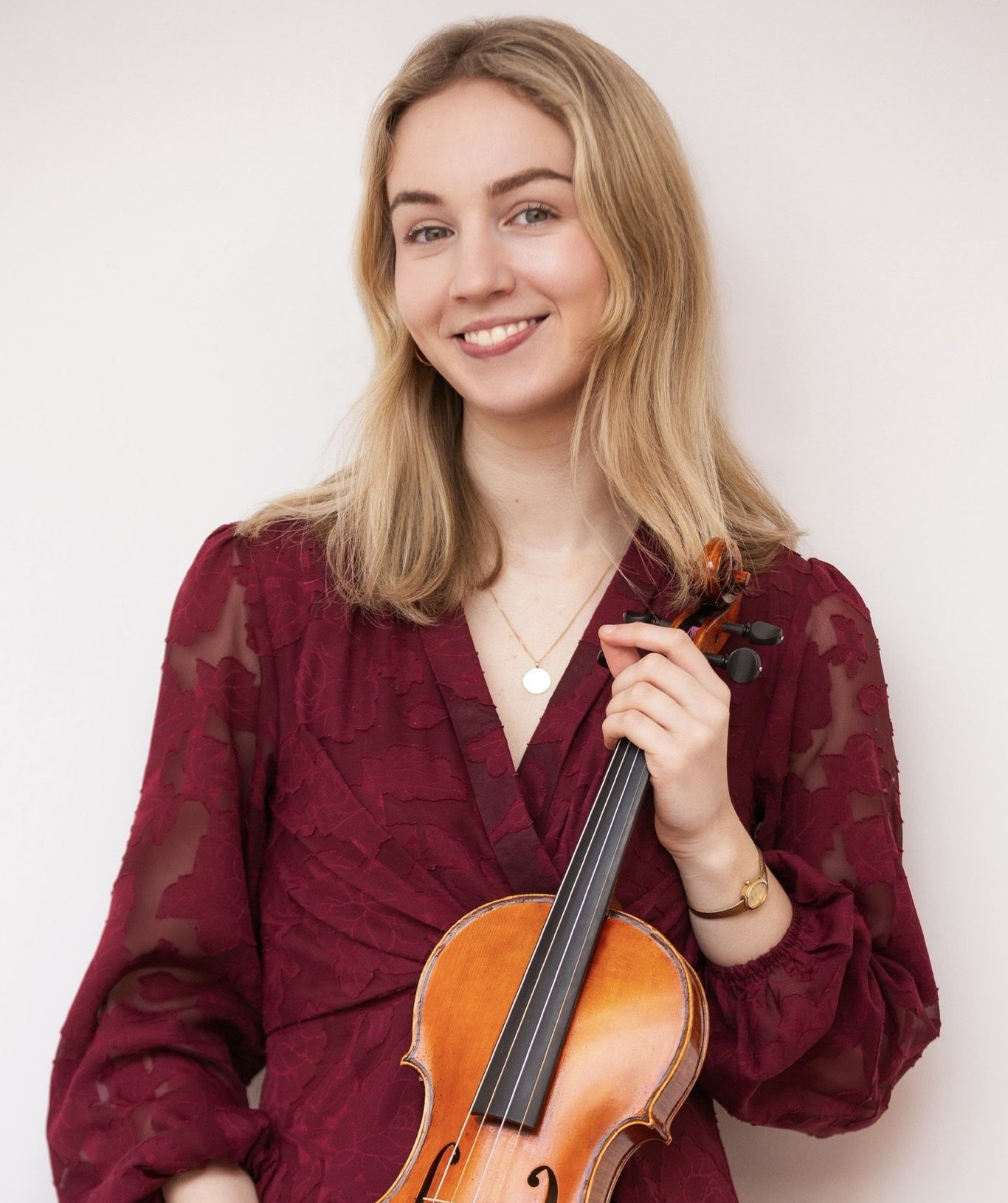 A young woman with blonde hair holding a violin, wearing a burgundy dress with a textured floral pattern, standing against a plain light background.