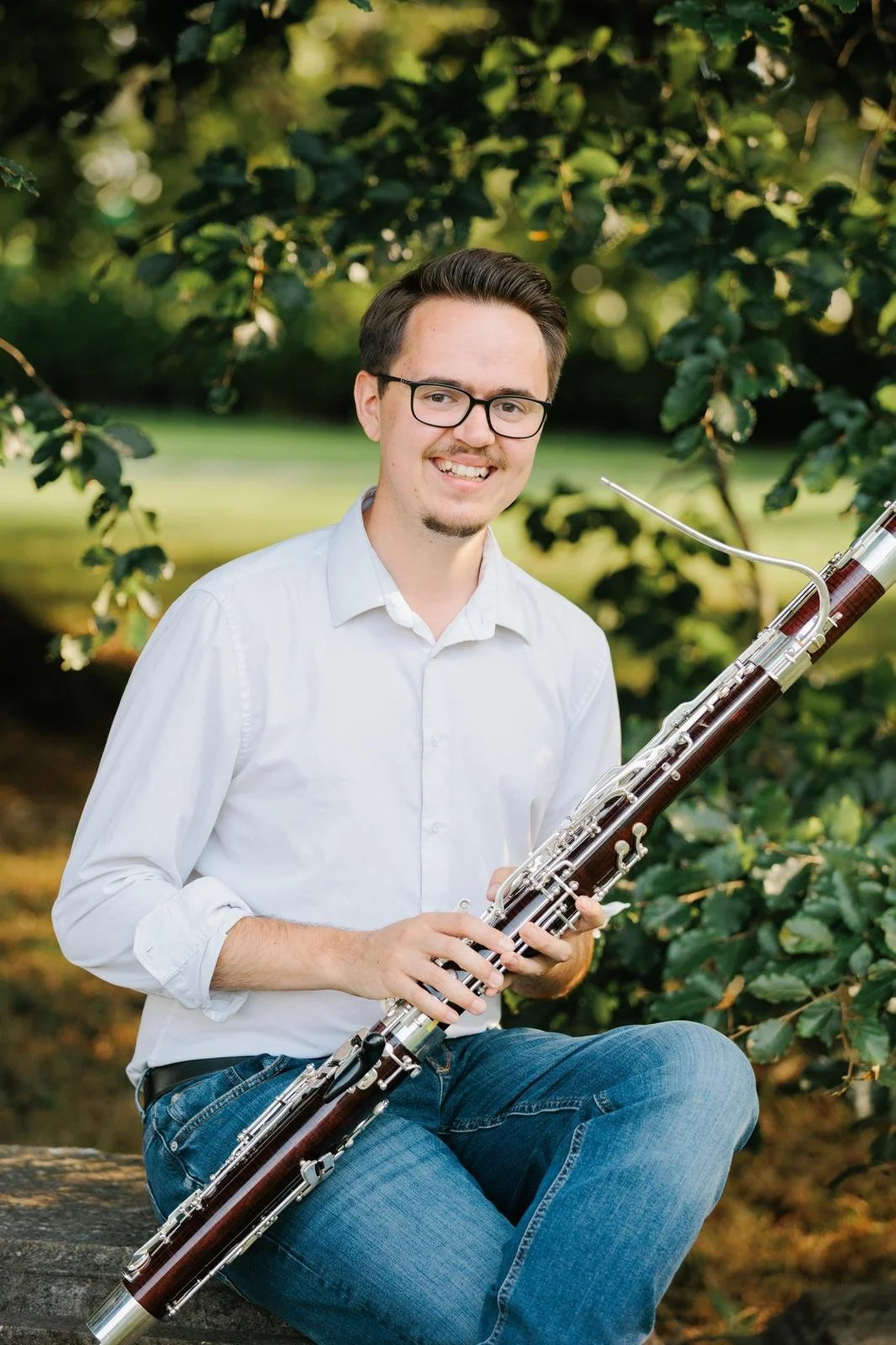Young man wearing glasses and a white shirt sitting outdoors on a log, holding a bassoon, with greenery in the background.