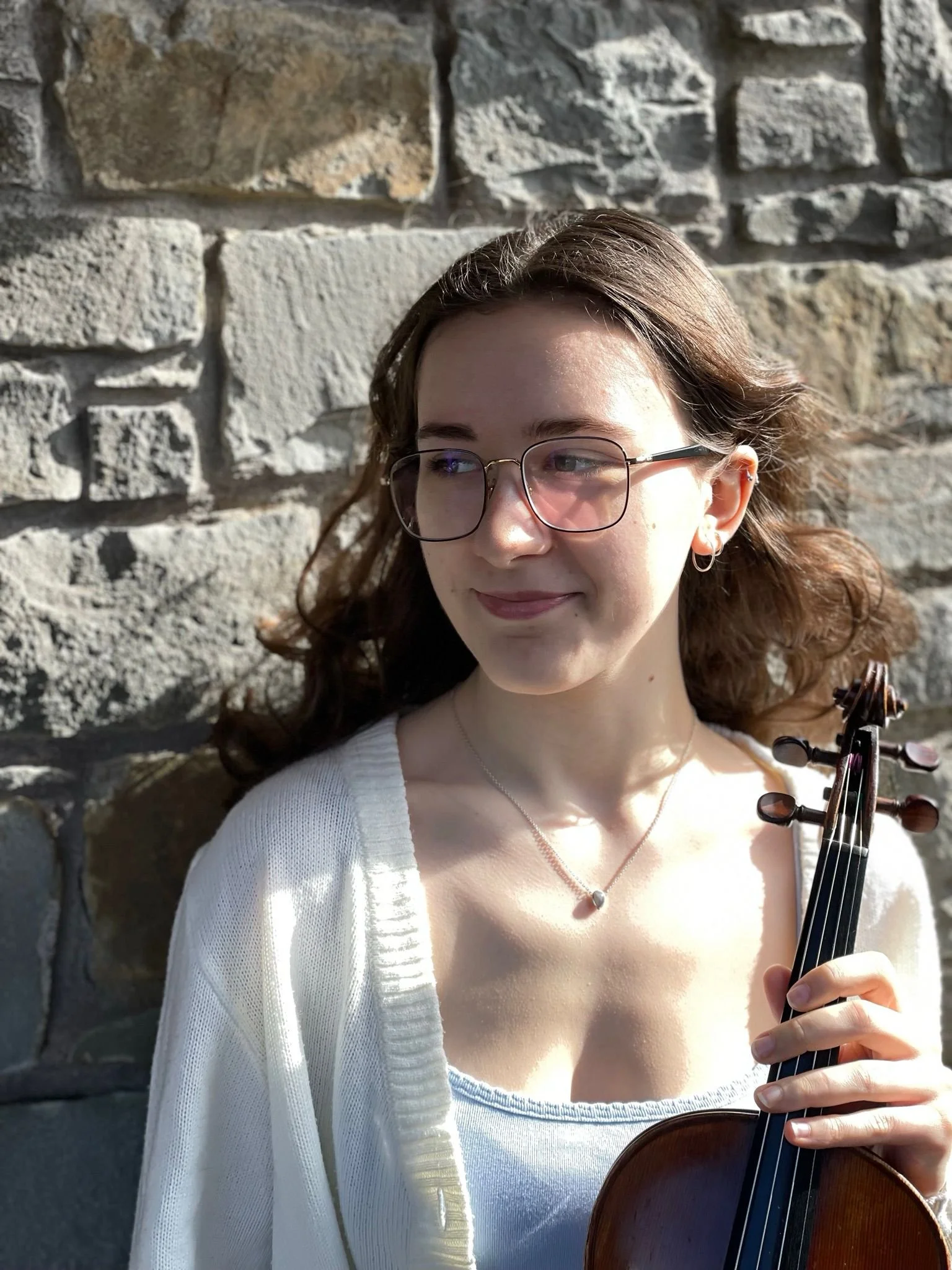 A young woman with brown curly hair, glasses, and earrings, holding a violin, stands in front of a stone wall, looking to her left.