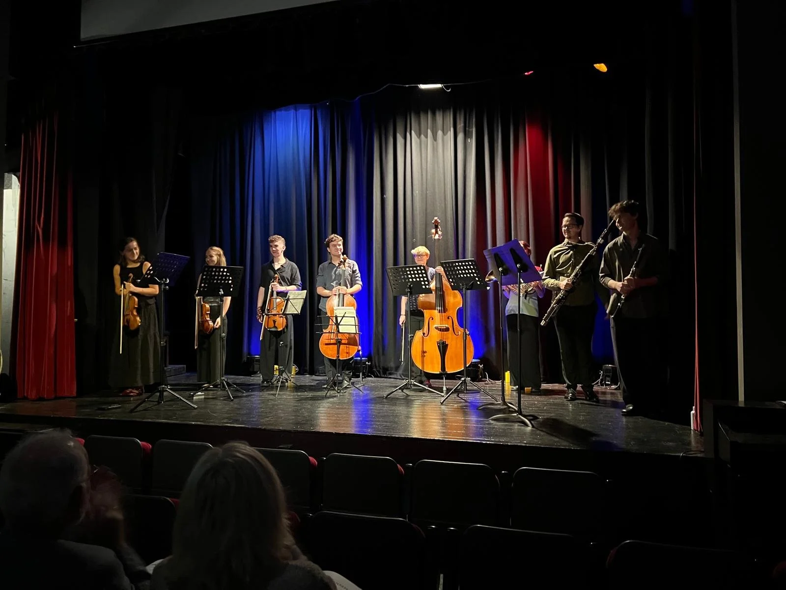 A group of eight young musicians on stage with musical instruments standing in line in front of a black curtain with coloured stage lighting, during a performance at Carnegie Arts Centre Kenmare.