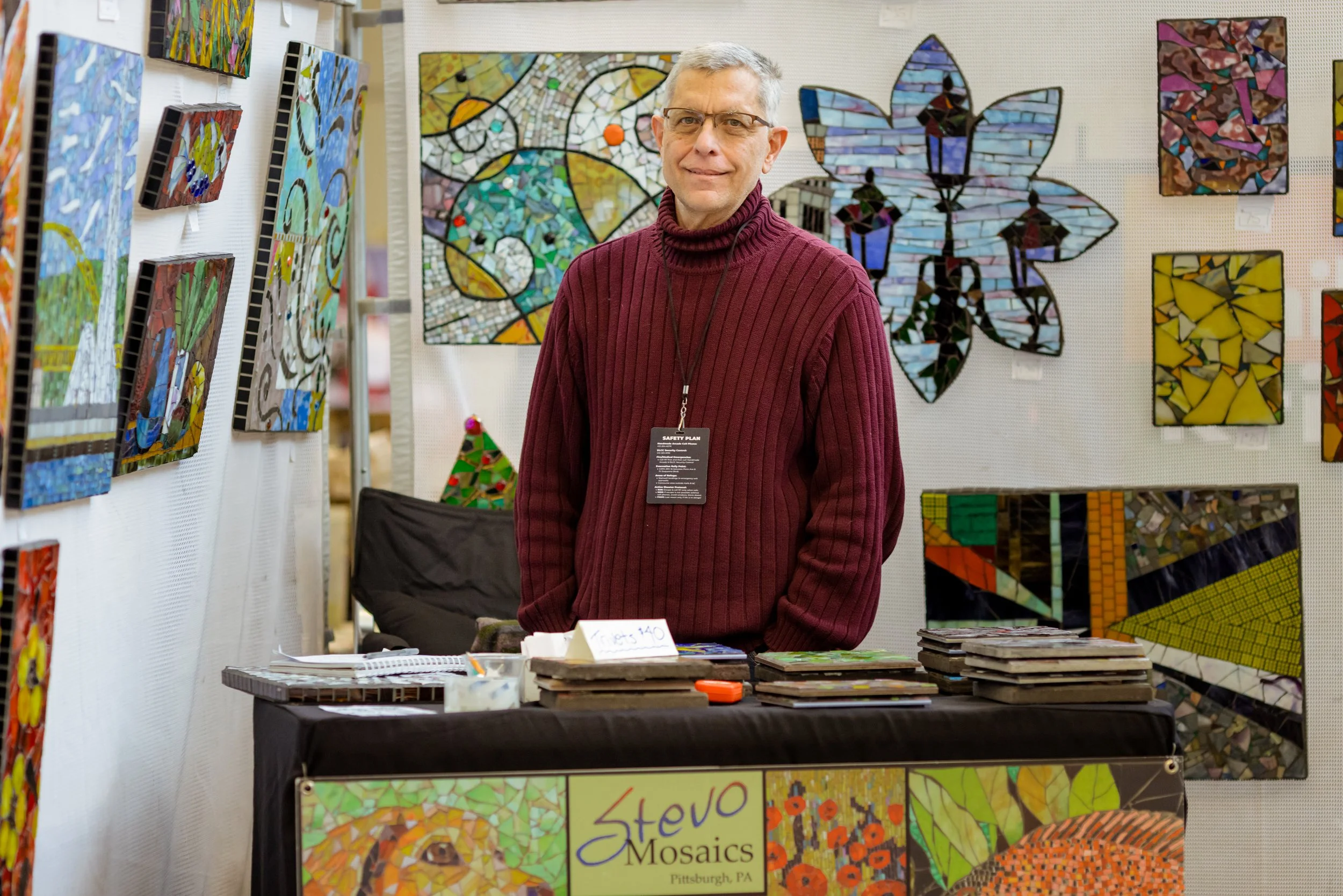 Male maker poses in front of his booth displaying vibrant handmade mosaic pieces.