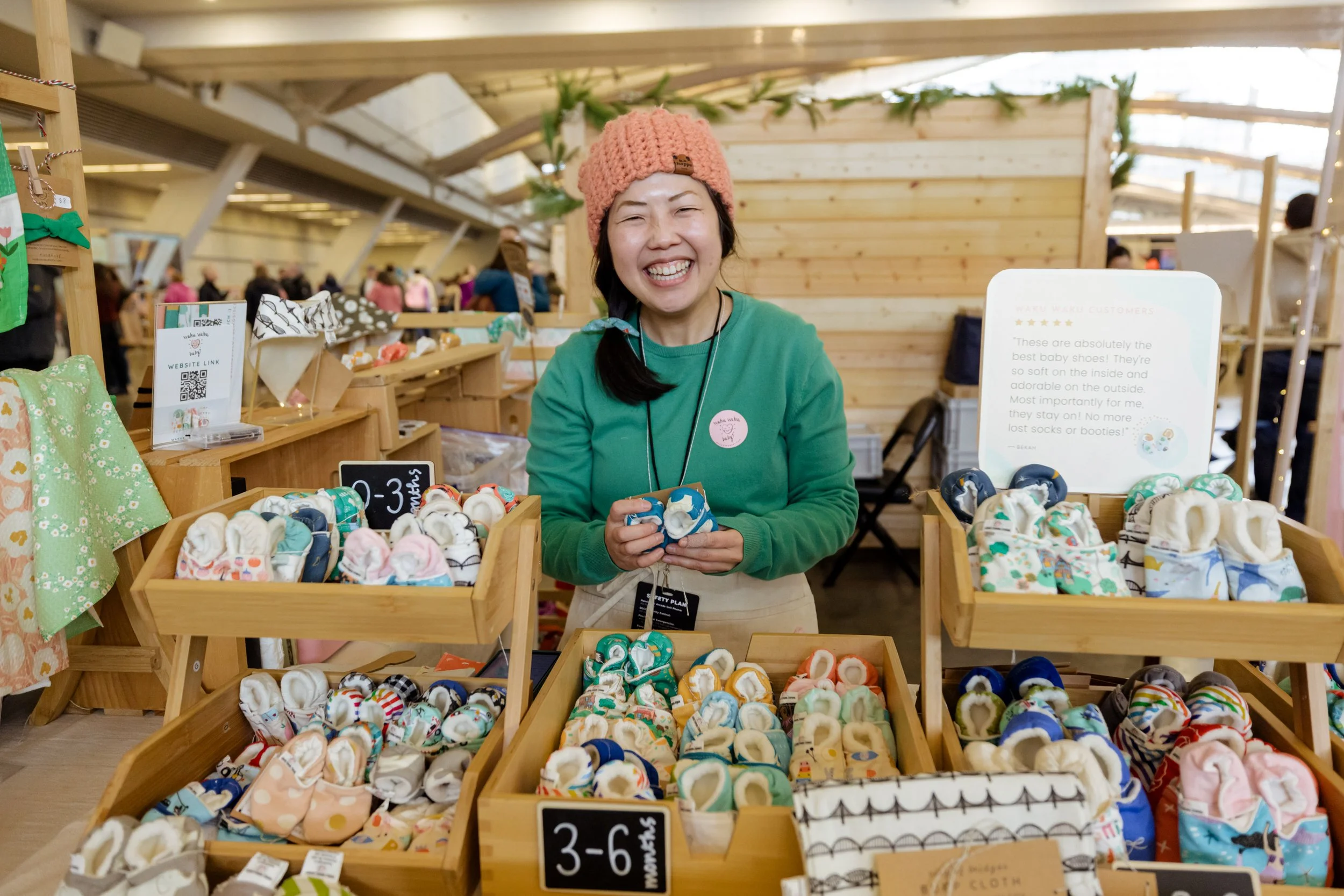 Close-up of a smiling maker holding handmade fabric baby shoes, standing behind bins of baby shoes labeled by age in months.