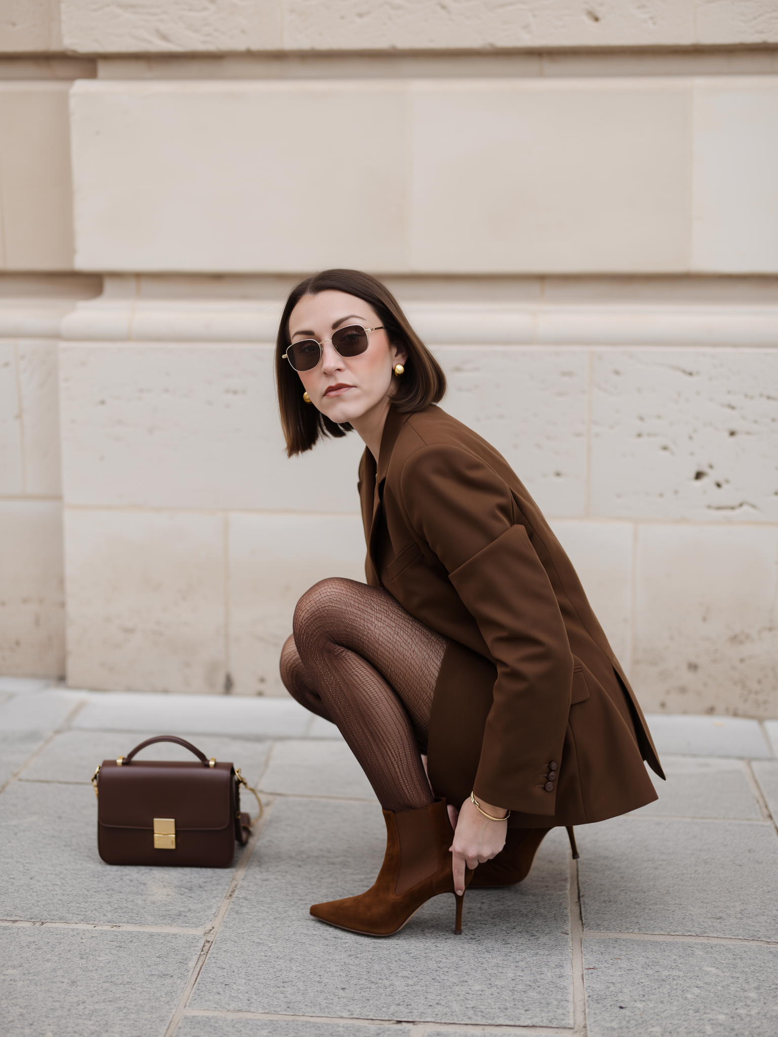 A woman with short brown hair, wearing sunglasses, a brown blazer, patterned tights, and brown high-heeled boots, crouches on a sidewalk next to a matching brown handbag in front of a beige stone building.