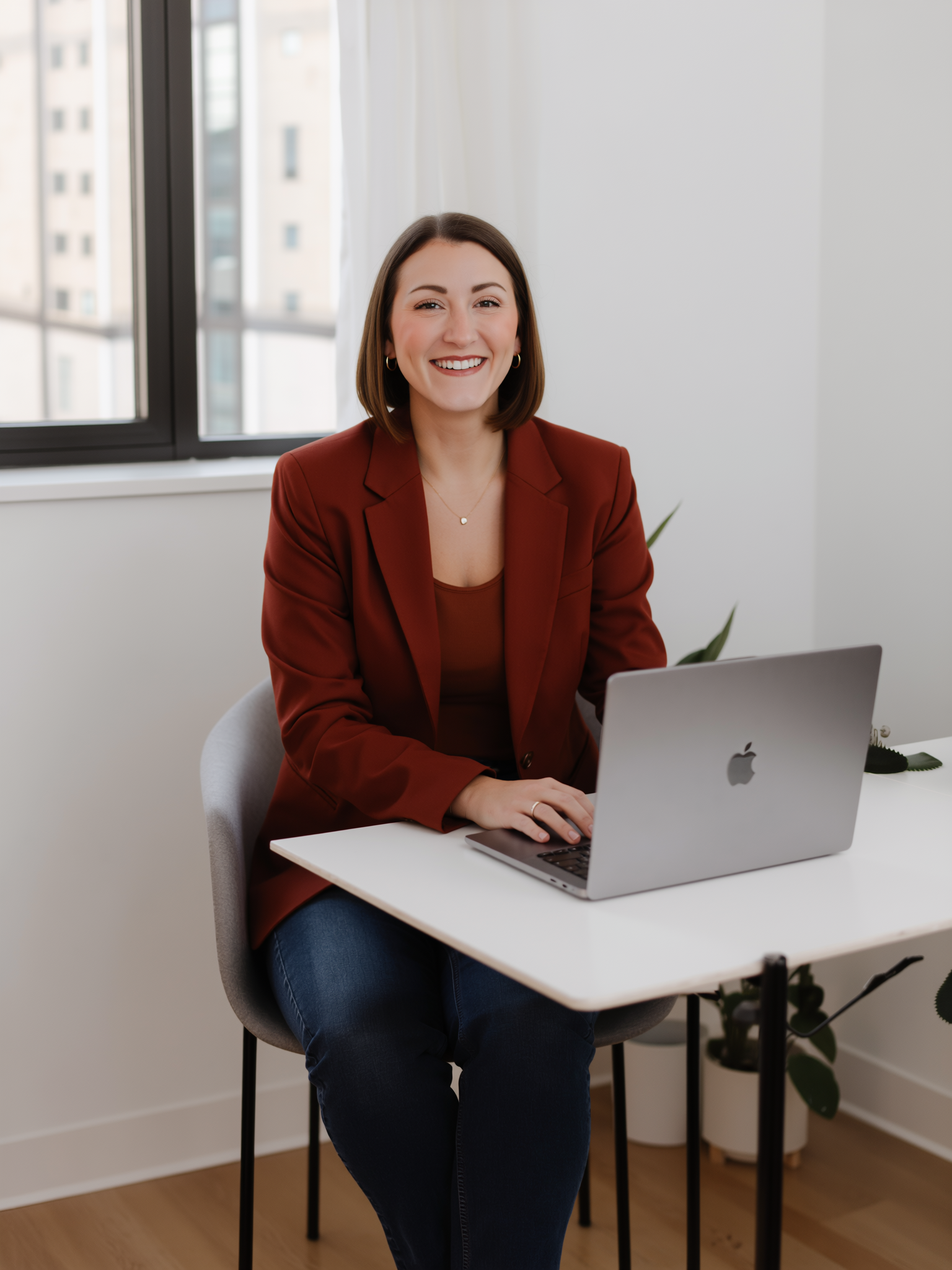 A woman with shoulder-length brown hair, smiling, wearing a red blazer, sitting at a white desk with a silver laptop in a bright office with large windows.
