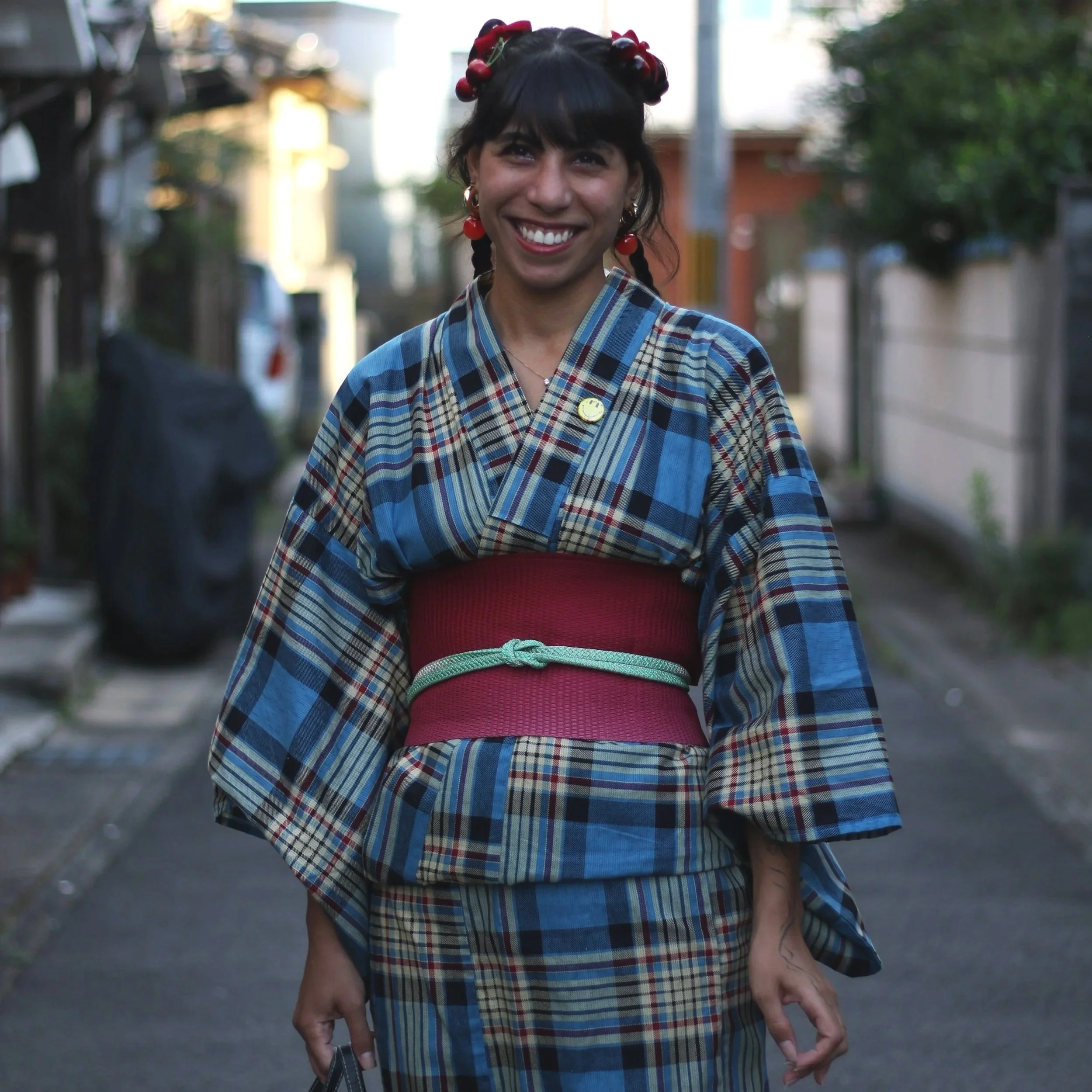A woman smiling in traditional Japanese kimono walking on a street with trees and houses in the background.