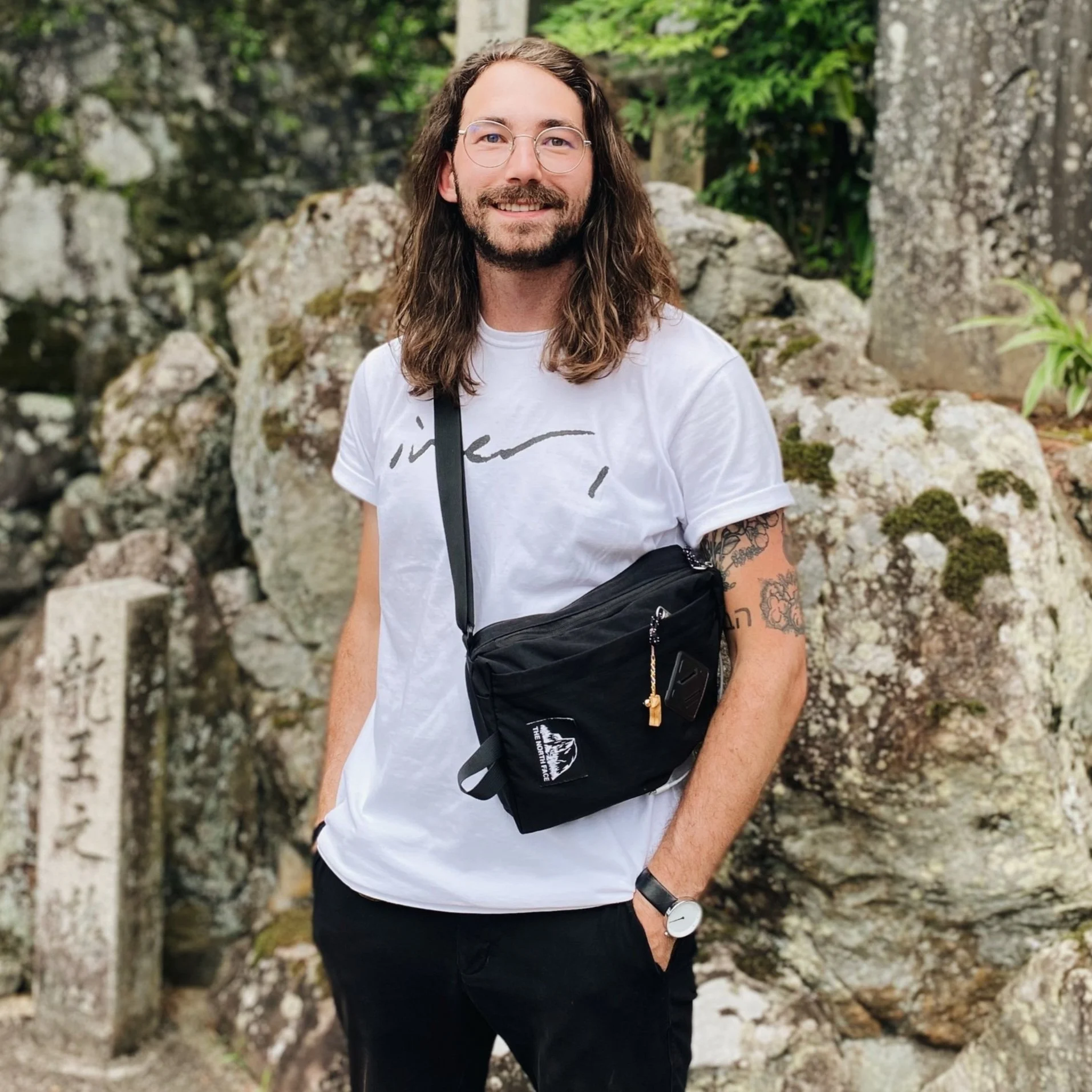A man with long brown hair, glasses, and a beard, wearing a white t-shirt with black text, black pants, a black bag across his chest, and a watch, standing outdoors in front of rocks and greenery.