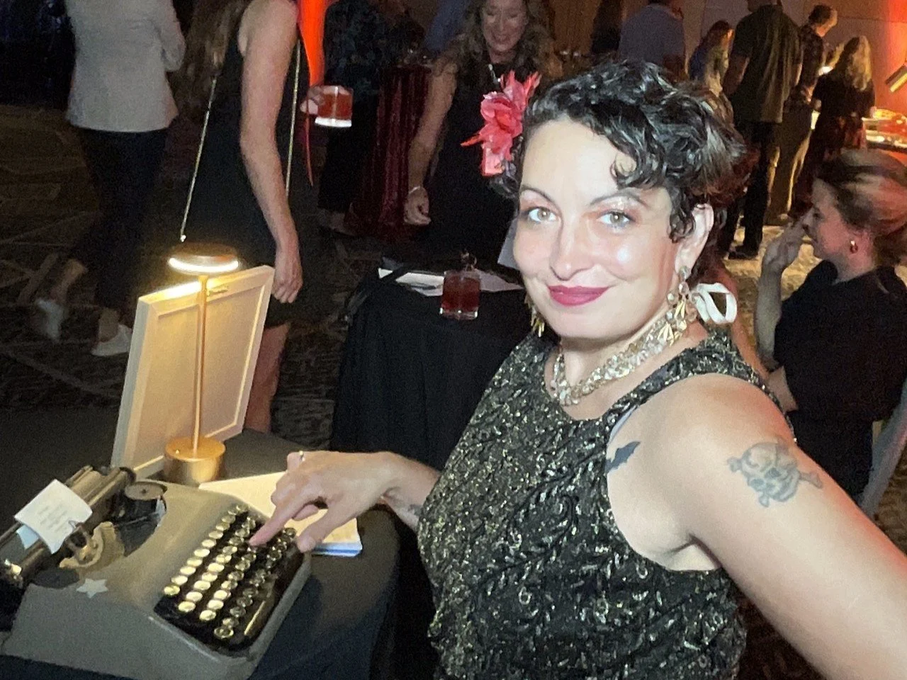 A woman with short curly hair, tattoos on her arm, and wearing a gold and black dress, smiling and typing on an old typewriter at a social event.