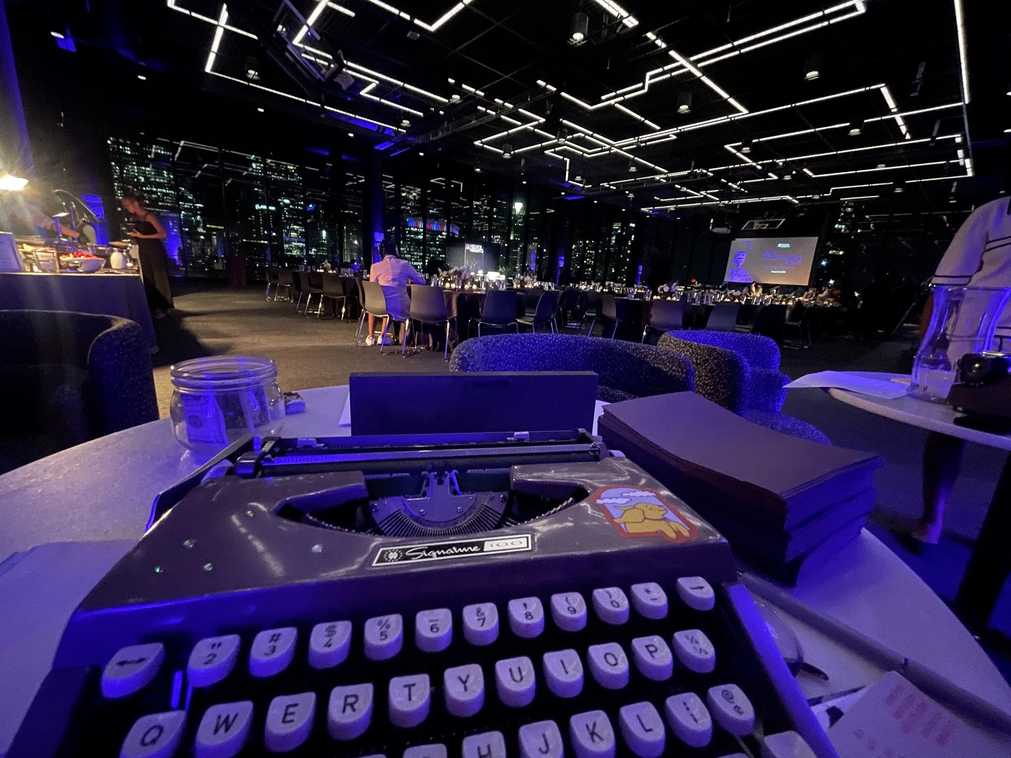 View of a typewriter at an event space in Chicago with large windows showing a city skyline at night, decorated with linear LED ceiling lights, with tables set for dining, and a DJ or music setup in the foreground.