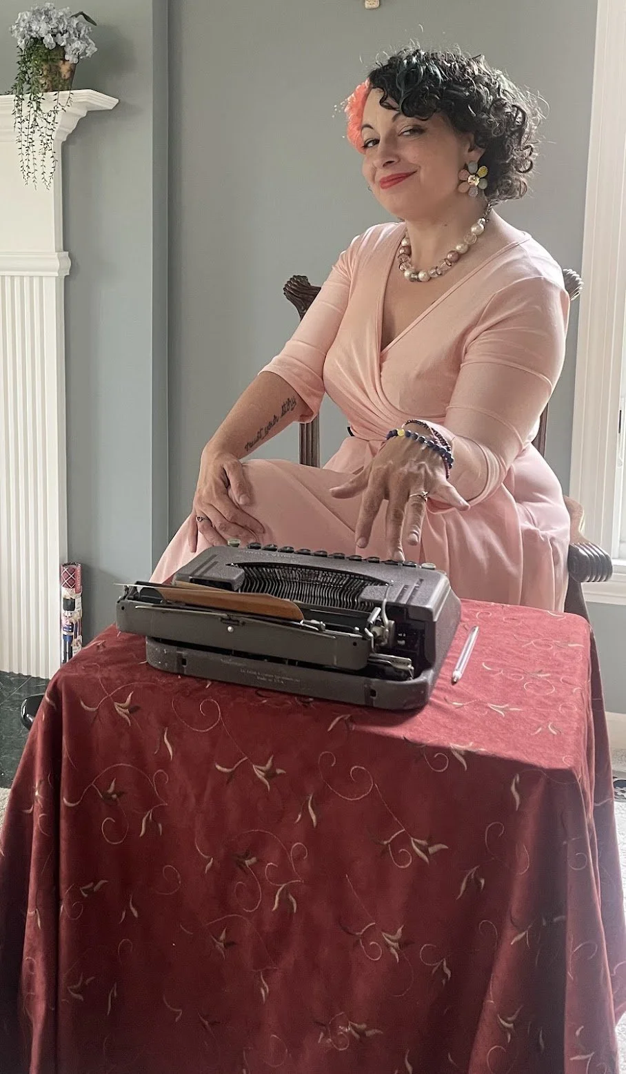 A woman dressed in a pink dress sitting at a table with a vintage typewriter. She is smiling, wearing colorful jewelry and strong makeup, with black and pink hair curl. There is a plant on a white shelf to her left and she is seated in front of a light gray wall.