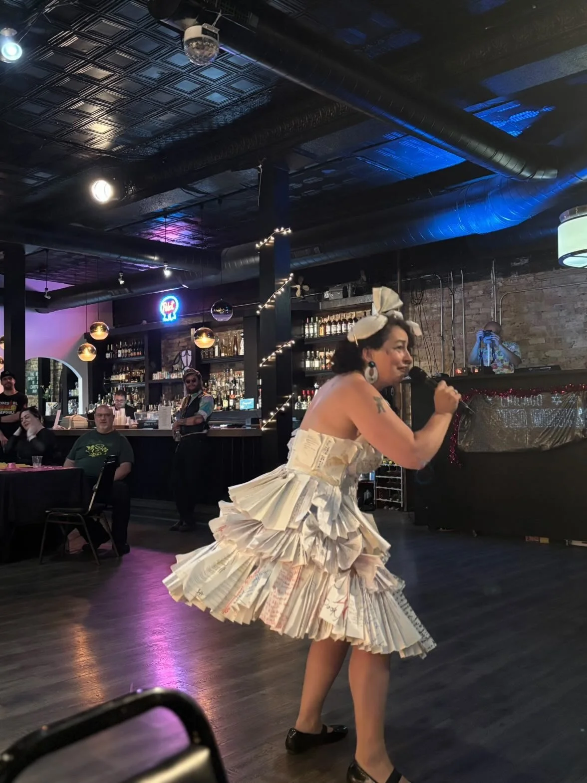 A woman performs on stage inside a bar or restaurant, wearing a dress made of folded paper or book pages, with a large bow on her head and holding a microphone. Several people are seated at tables and standing around, watching the performance. The ba
