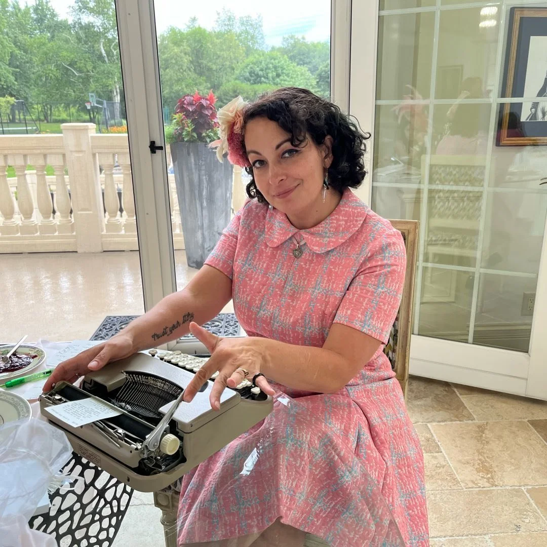 A woman sitting at a table with a vintage typewriter, wearing a pink patterned dress and a flower headband, smiling at the camera inside a room with large windows showing an outdoor patio and greenery.