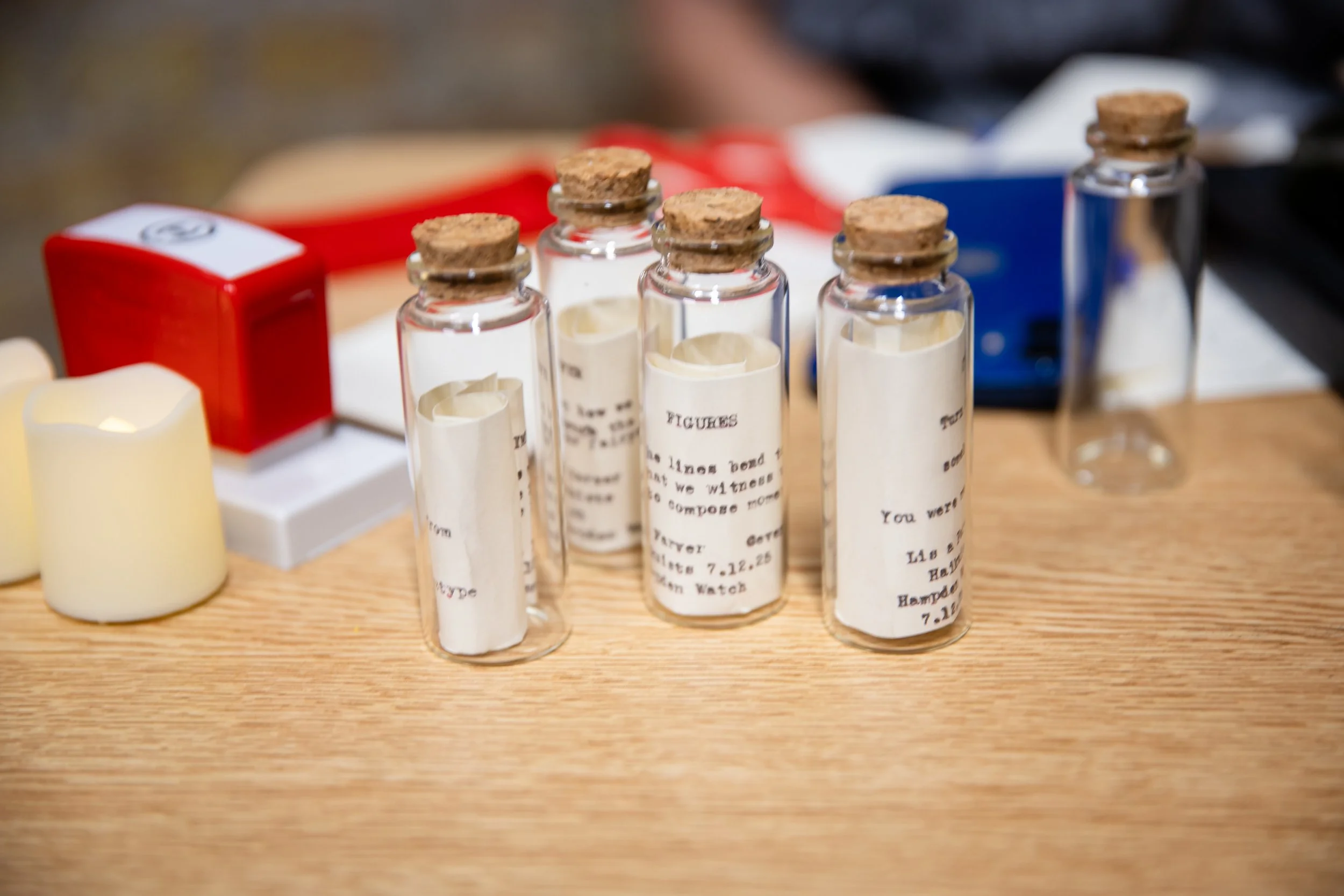 A close-up of small glass vials with cork stoppers, each labeled with handwritten notes, placed on a wooden surface, with a red box and a battery in the background.