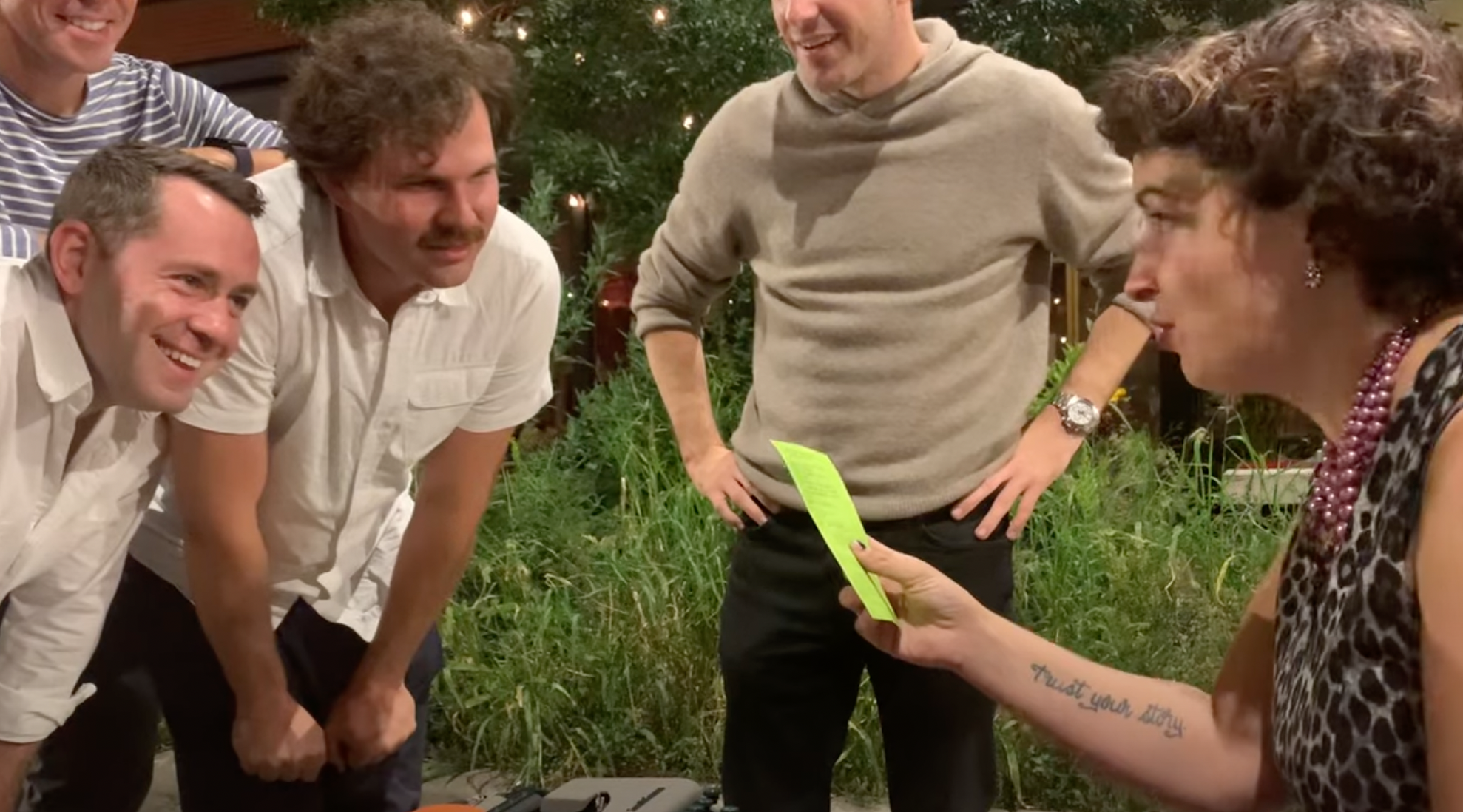 A group of five people gathered outdoors at night, engaged in a conversation. Three men are leaning forward, smiling, while a woman on the right holds a green paper, with a tattoo of text on her arm. In the background, there are trees with string lig