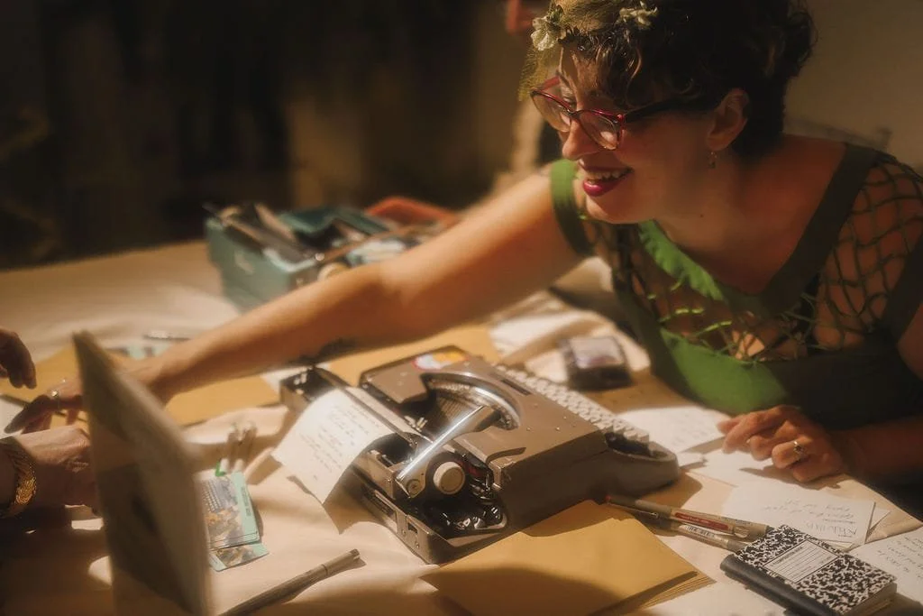 A typewriter poet with curly hair, glasses, and a floral headband is smiling and reaching across a table covered with papers, a vintage typewriter, a pen, and other office supplies.She is handing a typewriter poem to a guest at an event in Chicago.