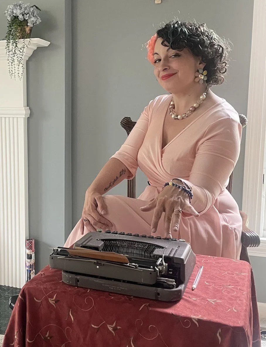 A woman with short curly dark hair with pink highlights, wearing a pink dress, pearl necklace, and floral earrings, sitting at a table with a vintage typewriter in front of her, smiling and pointing at it.