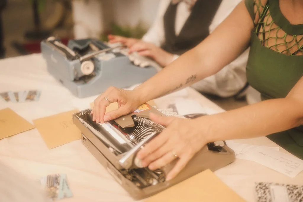 Two women are using vintage typewriters on a table with papers and small photographs, one wearing a green dress with a netted top and the other partially visible.
