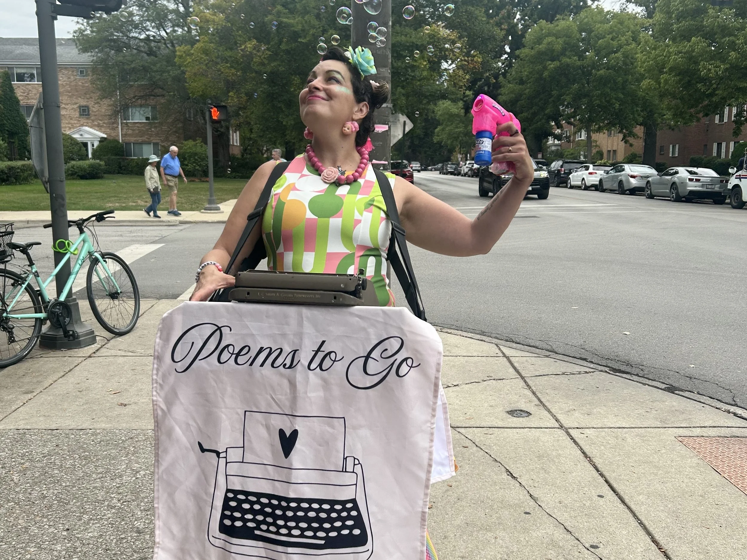 A woman with colorful accessories and makeup holding a bubble gun, standing outdoors at an event with a sign that reads 'Poems to Go', and people walking in the background.