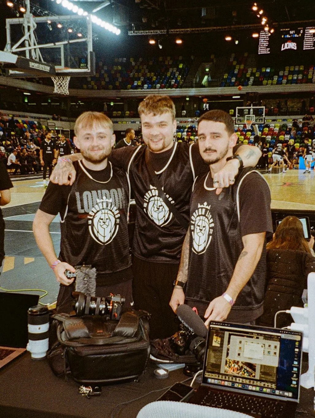 Three young men in black sports jerseys with a lion logo, smiling and posing together at a basketball game, in an indoor arena with a crowded background and colorful seats.