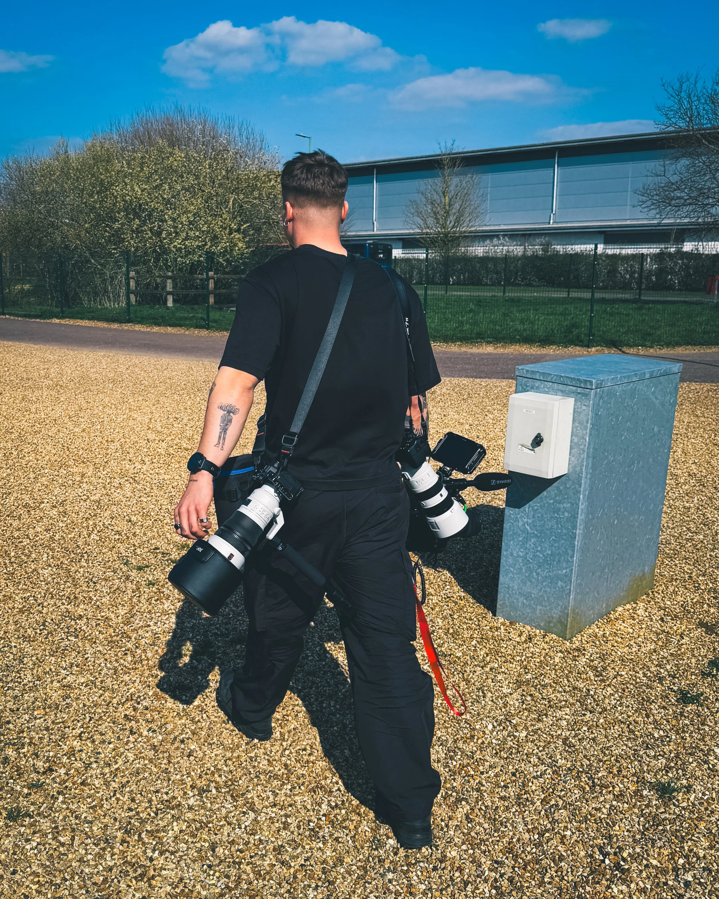 A man walking outdoors with multiple camera lenses hanging from his shoulders, wearing a black shirt and pants, with a tattoo on his left arm, near a metal utility box on a gravel surface under a blue sky.