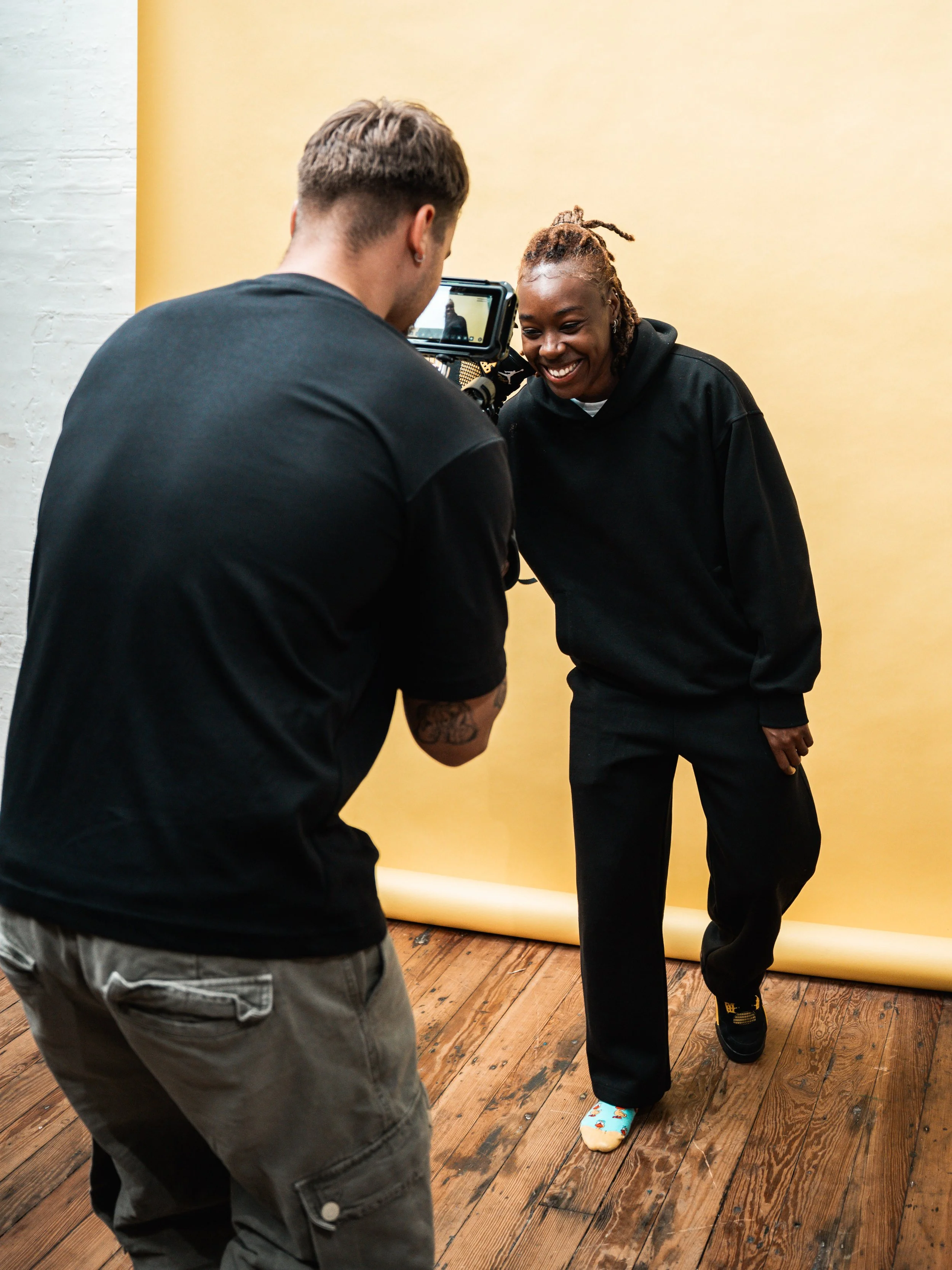 A woman is posing for a photo shoot while a man takes her picture against a yellow backdrop.