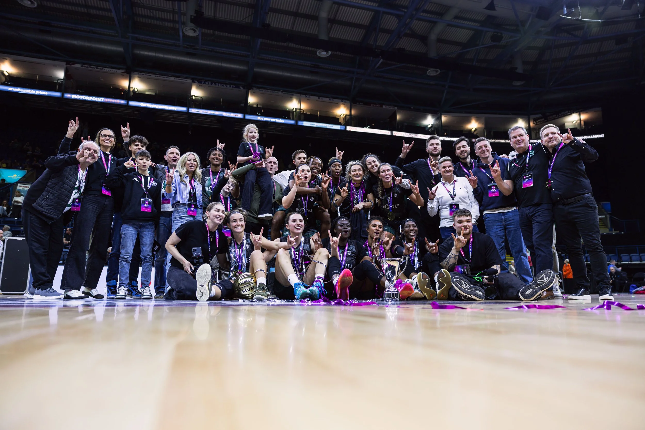 Group of women athletes and supporters celebrating victory on an indoor basketball court, wearing medals and holding trophies, with a crowd and arena in the background.