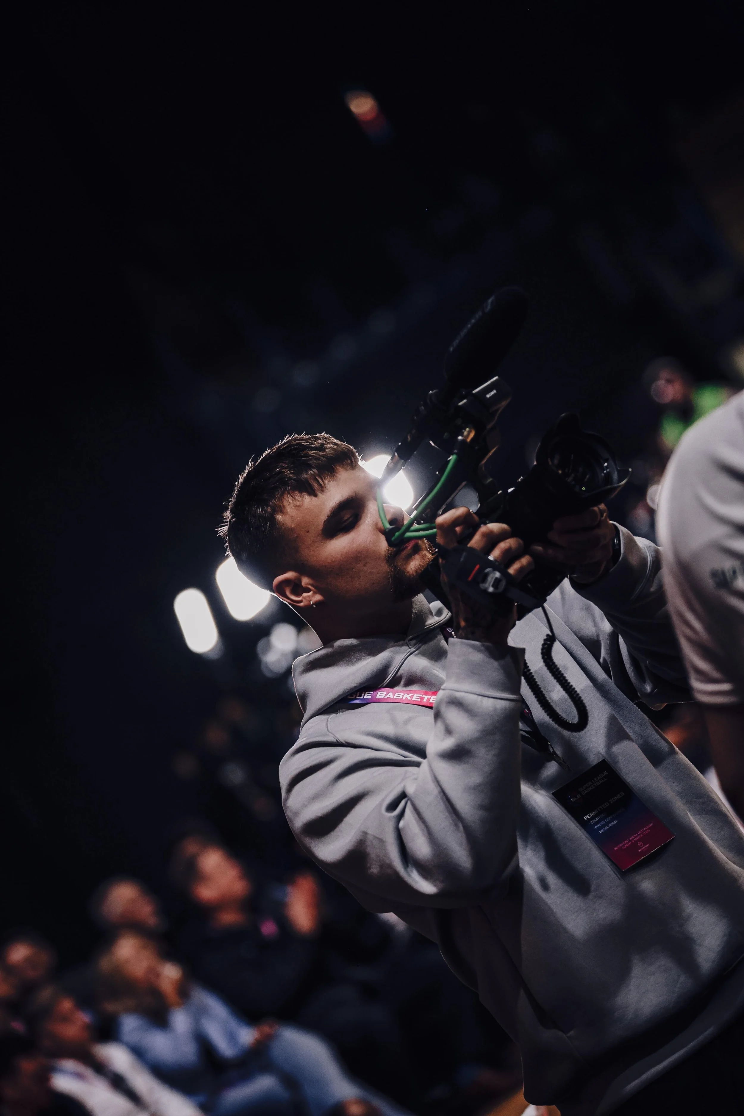 A man operating a professional video camera at an indoor event, with focused expression and a crowd in the background.