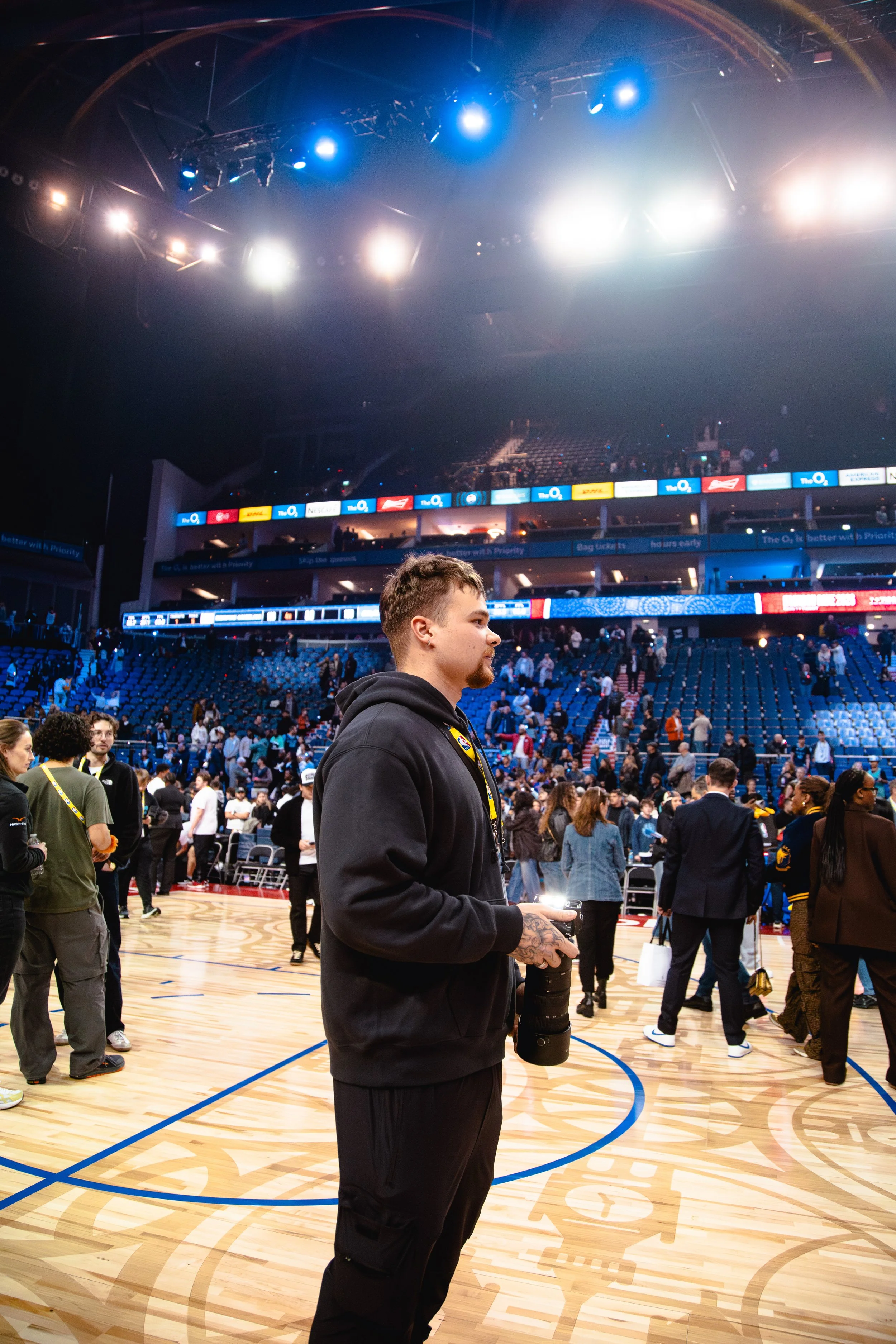 Young man with tattoos wearing black hoodie holding a camera inside a sports arena with a crowd, bright overhead lights, and blue seating.