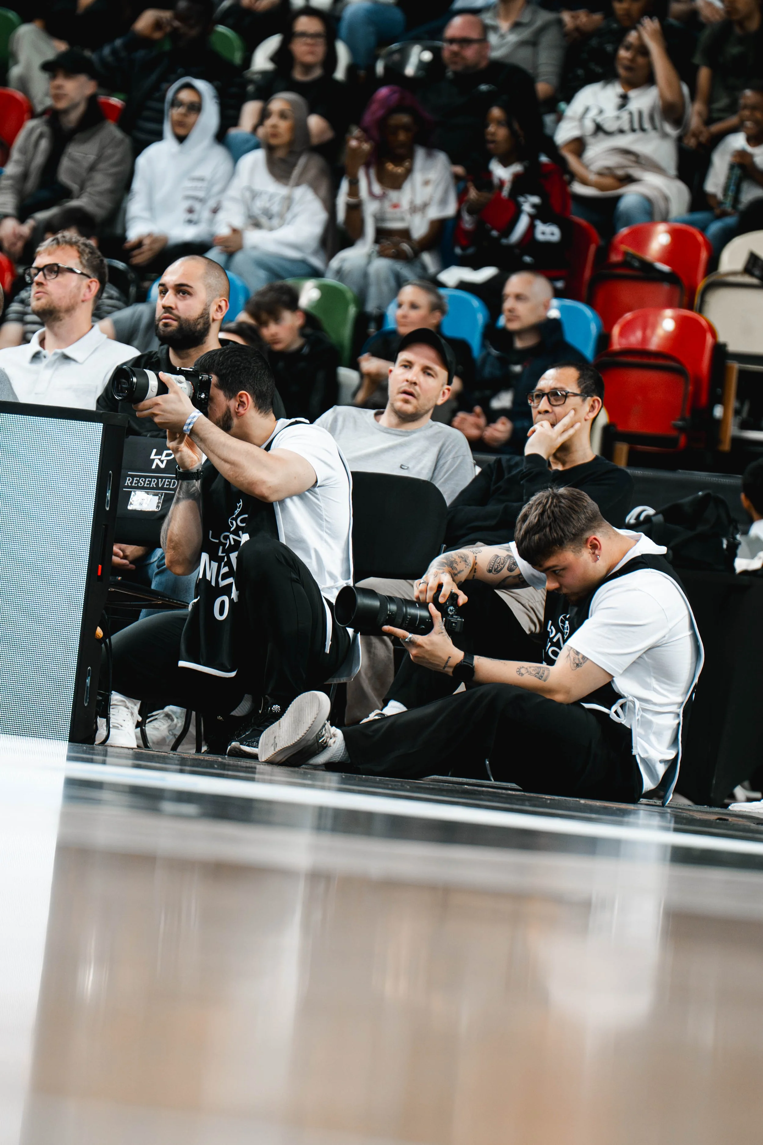 Photographers sitting on the floor with cameras, capturing an event in front of them, surrounded by an audience seated in colorful chairs.