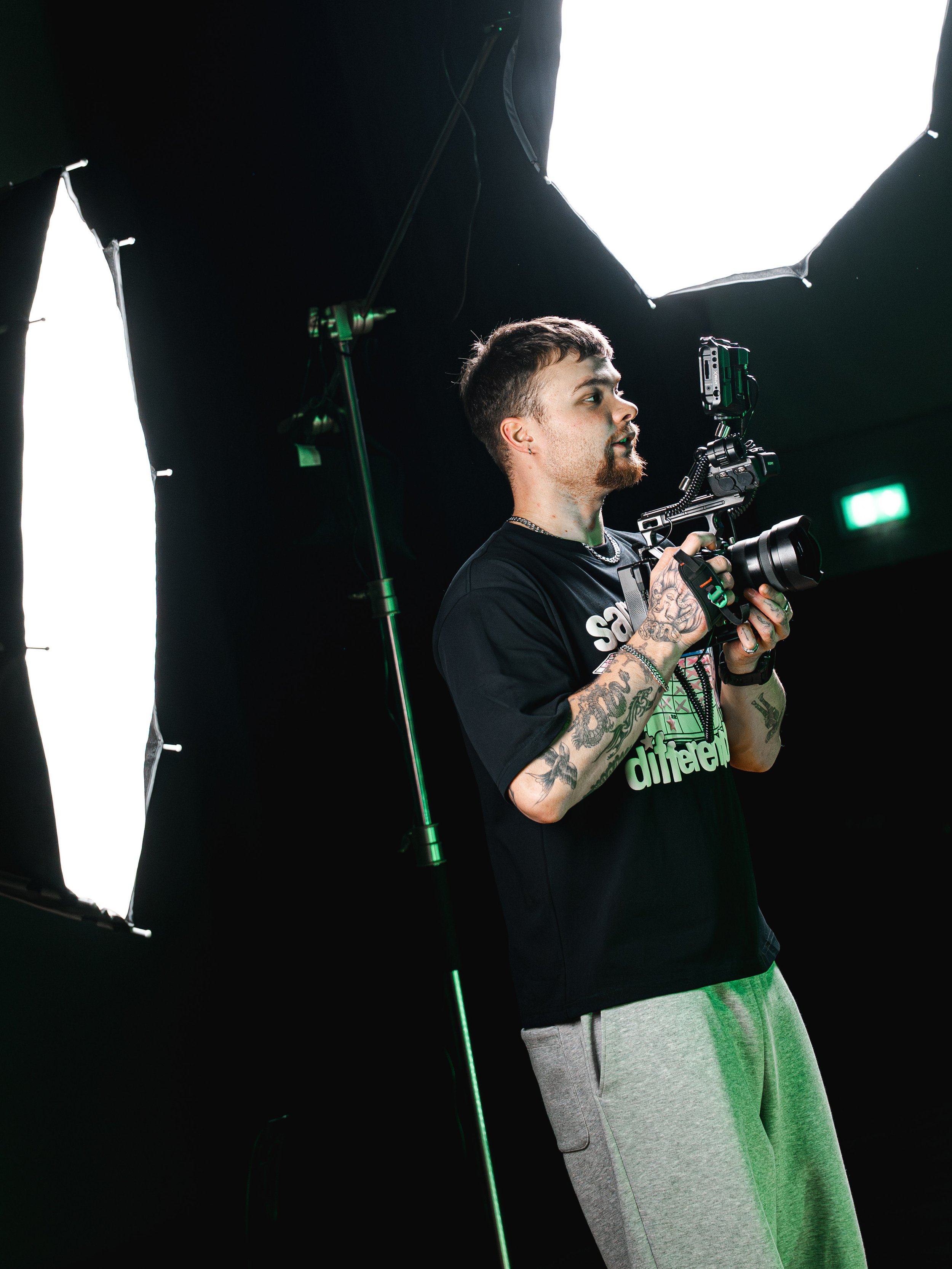 A man with tattoos on his arms holds a professional camera on a stabilizer, standing in front of studio lights.