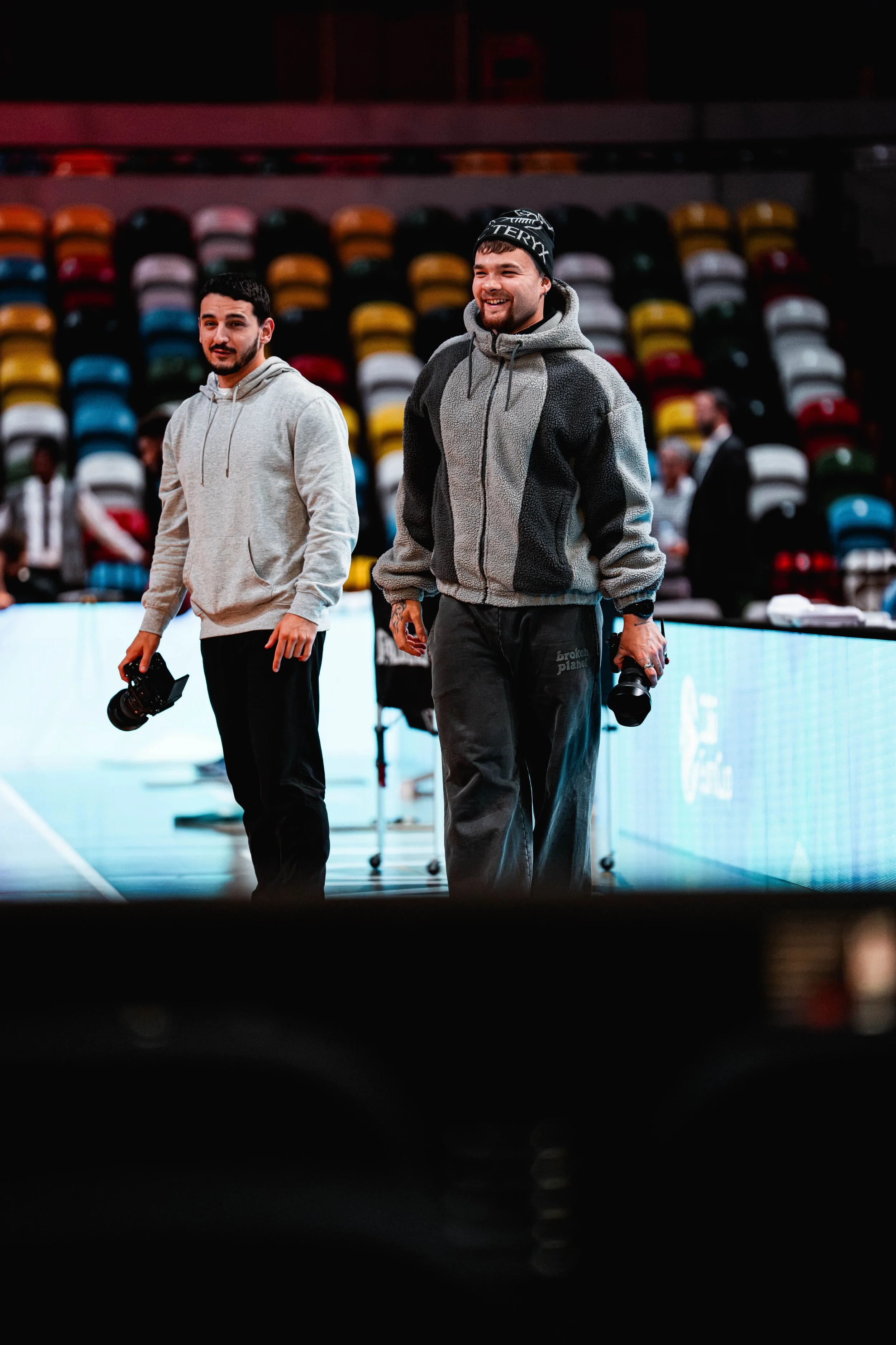 Two men holding cameras in an indoor arena with multicolored empty seats in the background.
