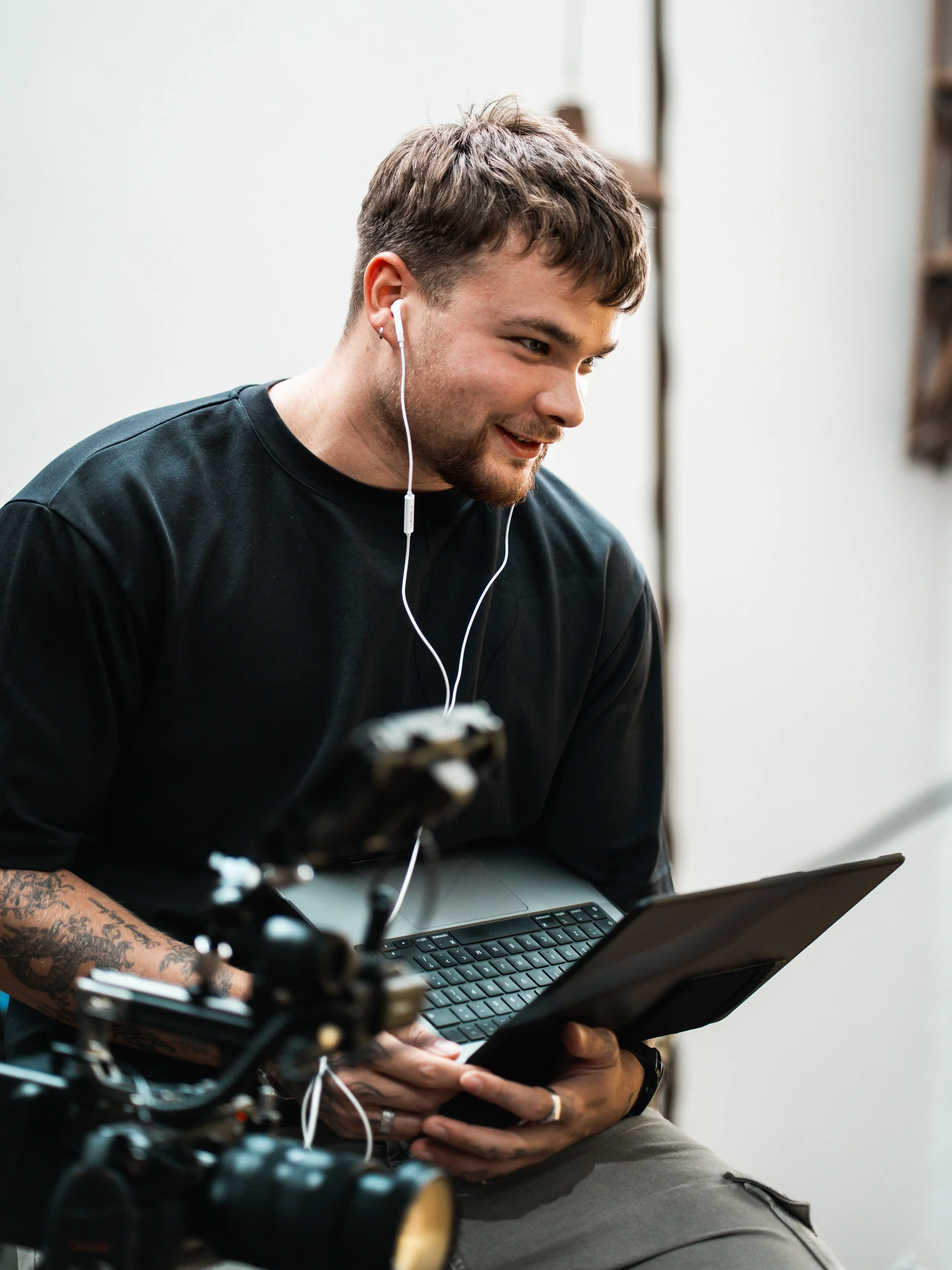 Young man with tattoos wearing a black t-shirt using a tablet and earbuds, sitting near a camera, in a bright indoor space.
