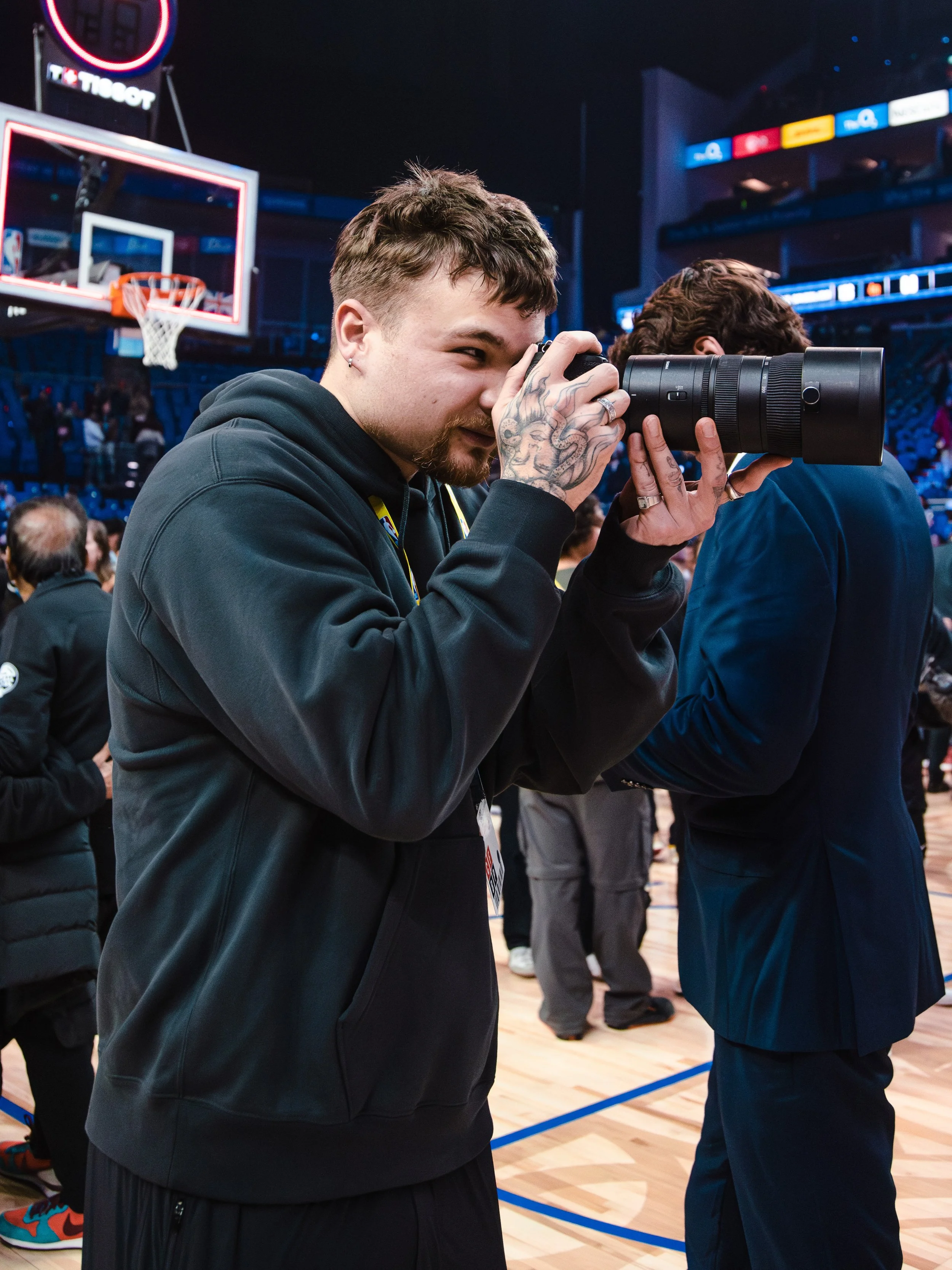 A man taking a photo with a professional camera in a sports arena, basketball hoop visible in the background, many people present in the venue.