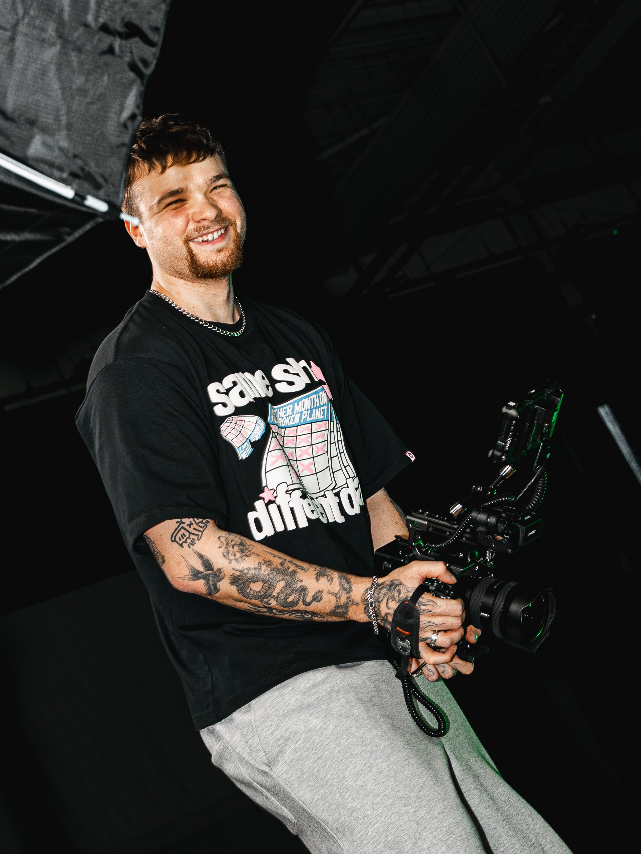 A young man with tattoos, a beard, and short brown hair holding a professional camera, smiling and wearing a black t-shirt with graphic text and gray sweatpants, standing in a dark studio setting.