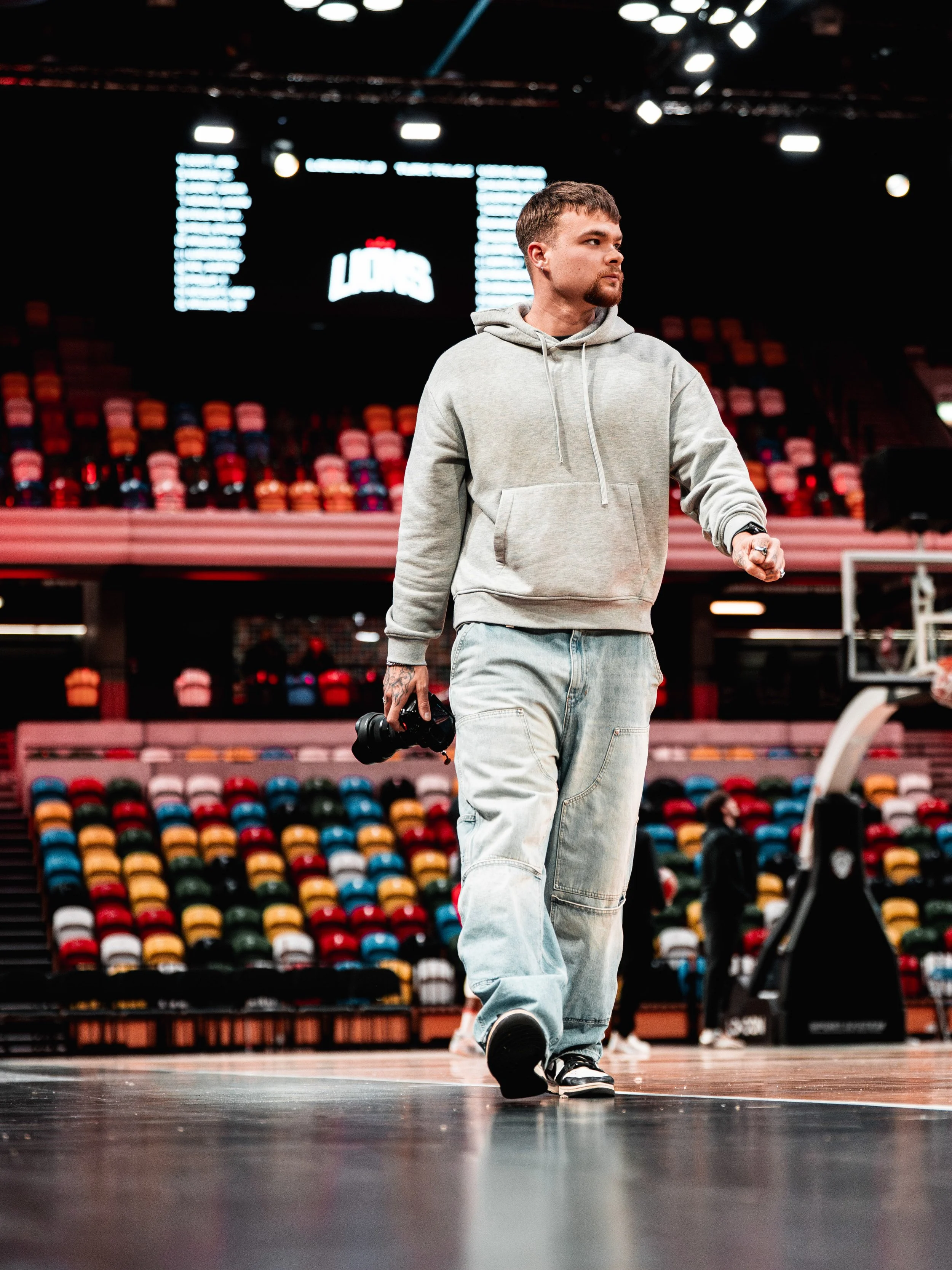 A man in a gray hoodie and light jeans walking on a basketball court, holding a camera, in an indoor arena with colorful seats.