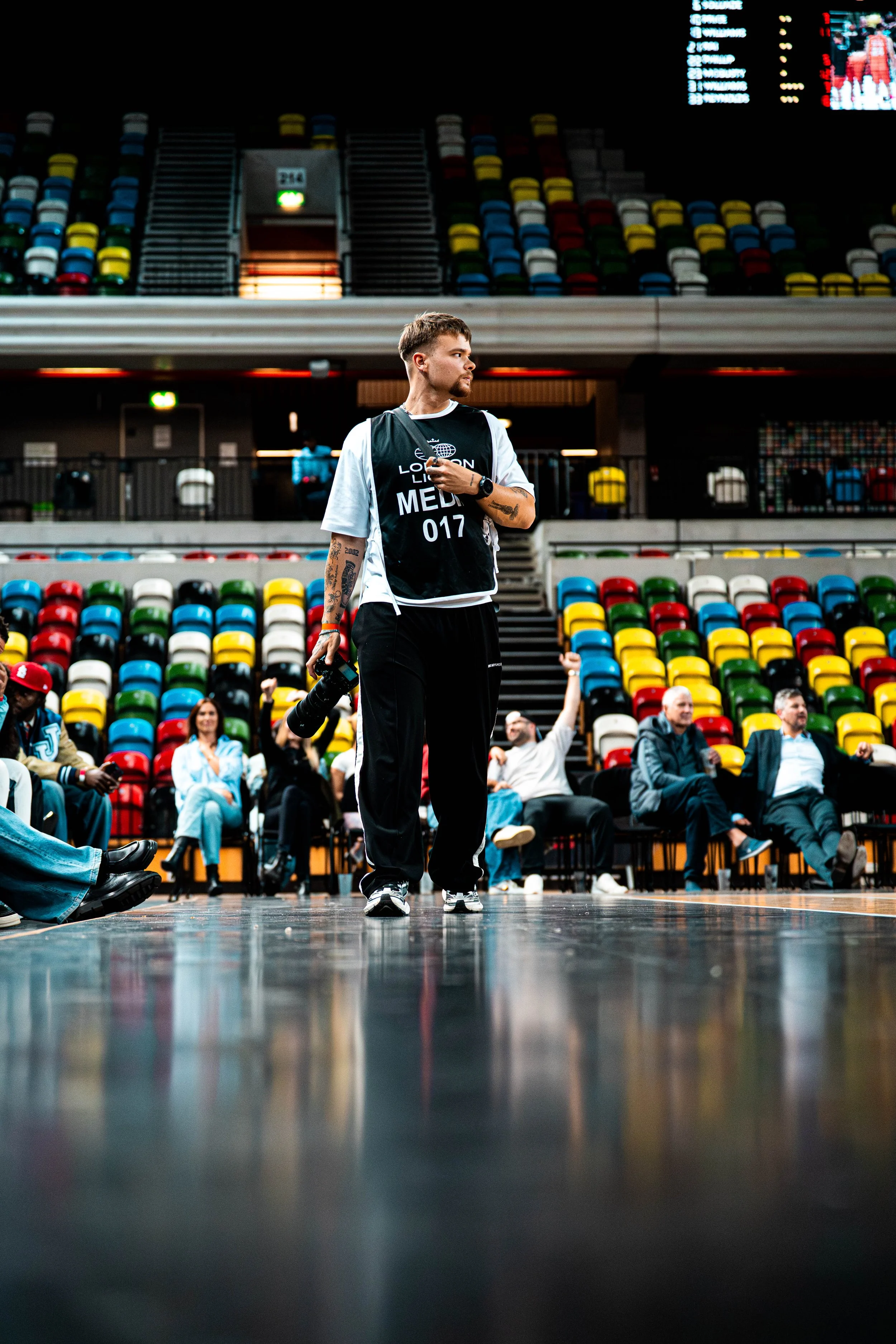 A young man with tattoos and athletic clothing standing alone in an empty sports arena, holding a camera and a walkie-talkie, with colorful seats and seated spectators in the background.