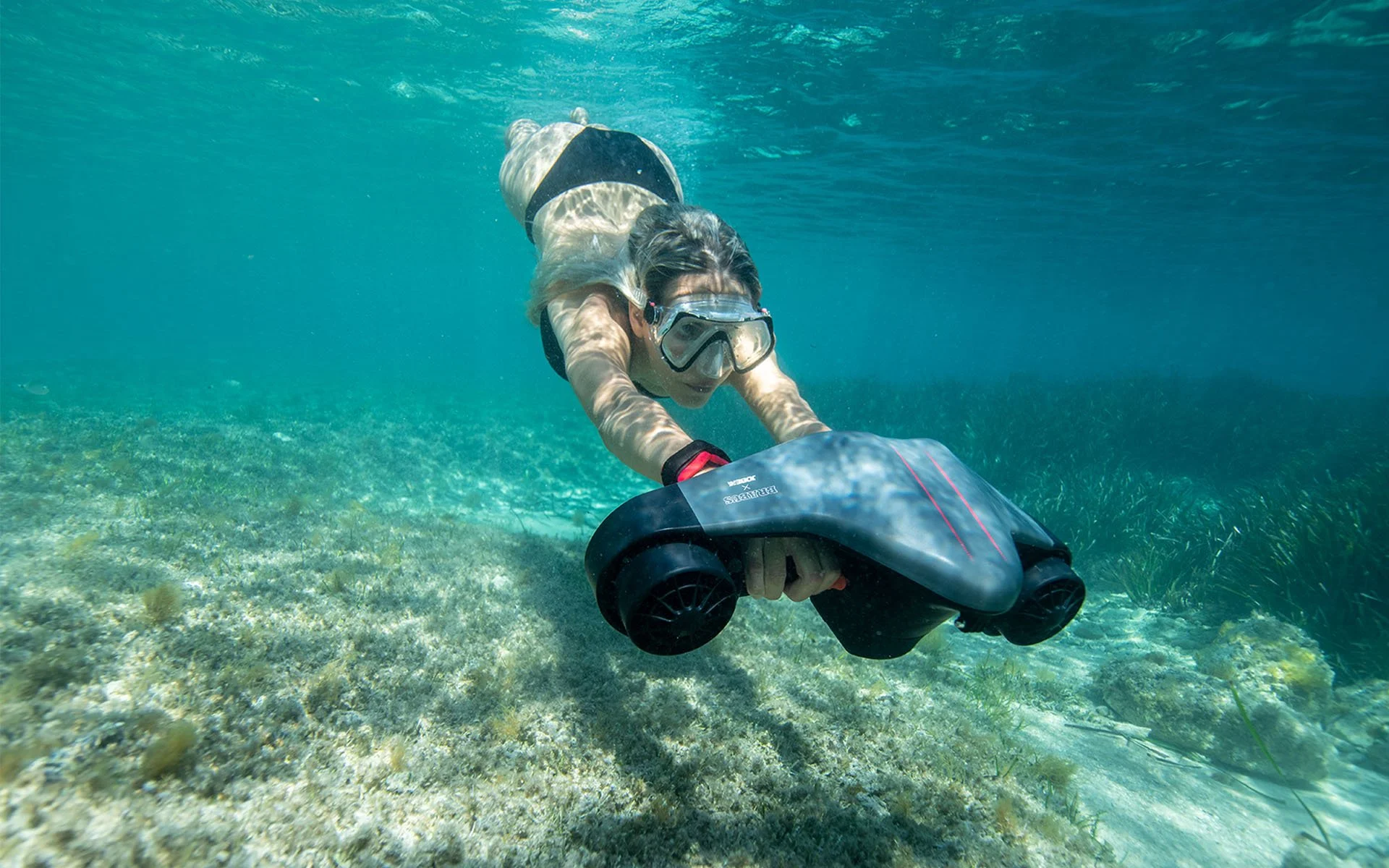Woman snorkeling underwater, reaching towards the sea floor with a waterproof camera, wearing a snorkel mask and swimming suit using a seascooter.