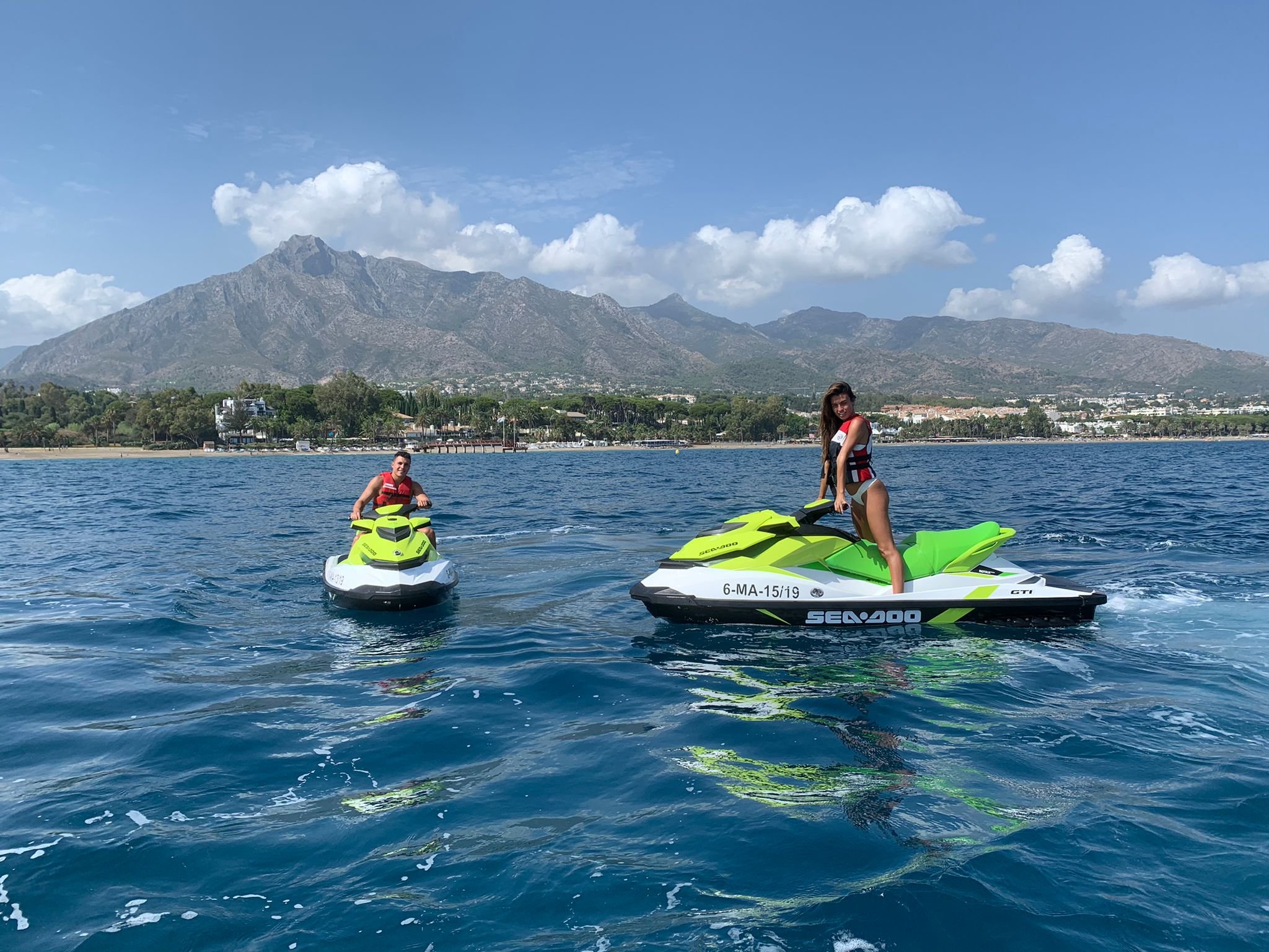 Beautiful woman riding and chilling on neon yellow and green jet skis on a large body of water with a mountainous landscape and some buildings in the background. Clear blue sky with a few clouds.