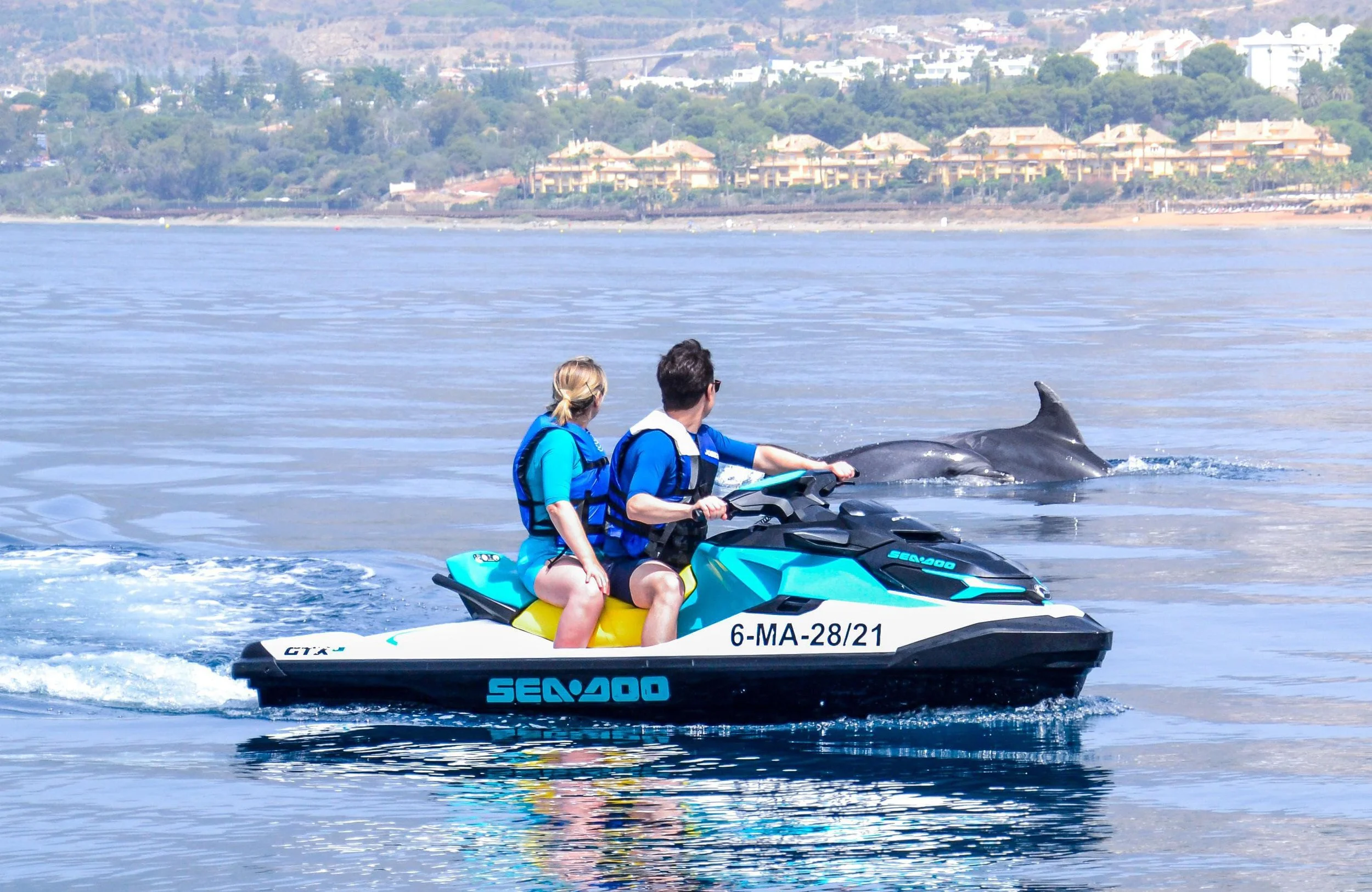 Two people riding a jet ski with a dolphin surfacing nearby on a calm body of water, with a shoreline and buildings in the background.