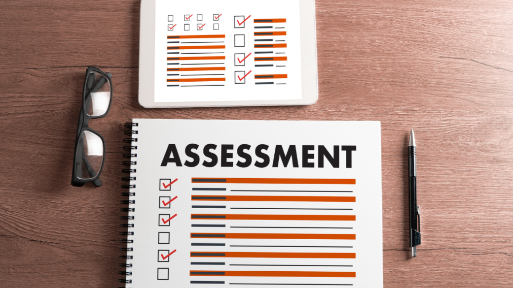 An assessment document with checked boxes, a tablet displaying check marks, a pair of glasses, and a pen on a wooden surface.