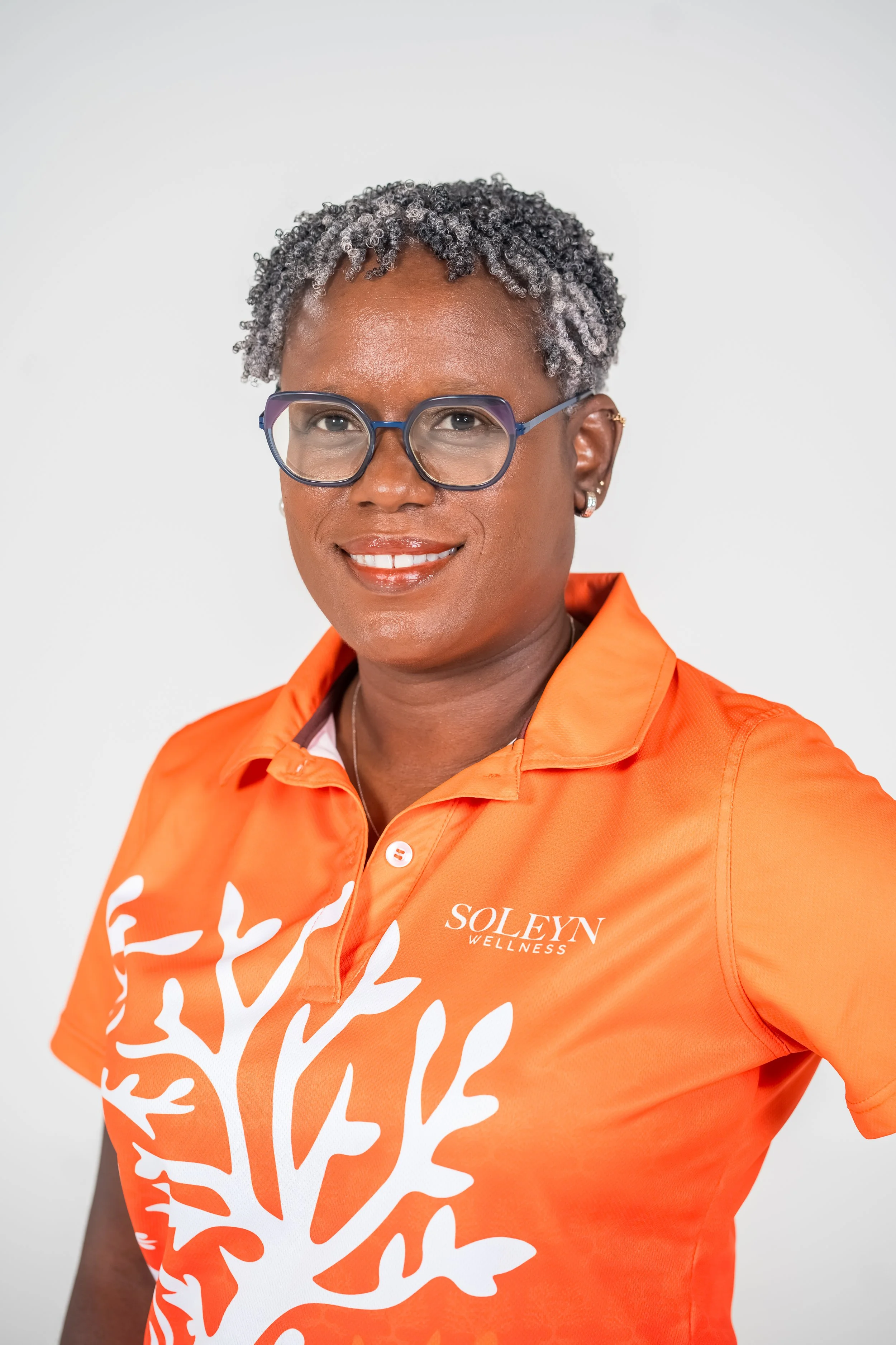 A woman with short, curly gray hair wearing glasses and an orange shirt with the text 'SOLEYN WELLNESS' and a white tree graphic, posing against a plain white background.