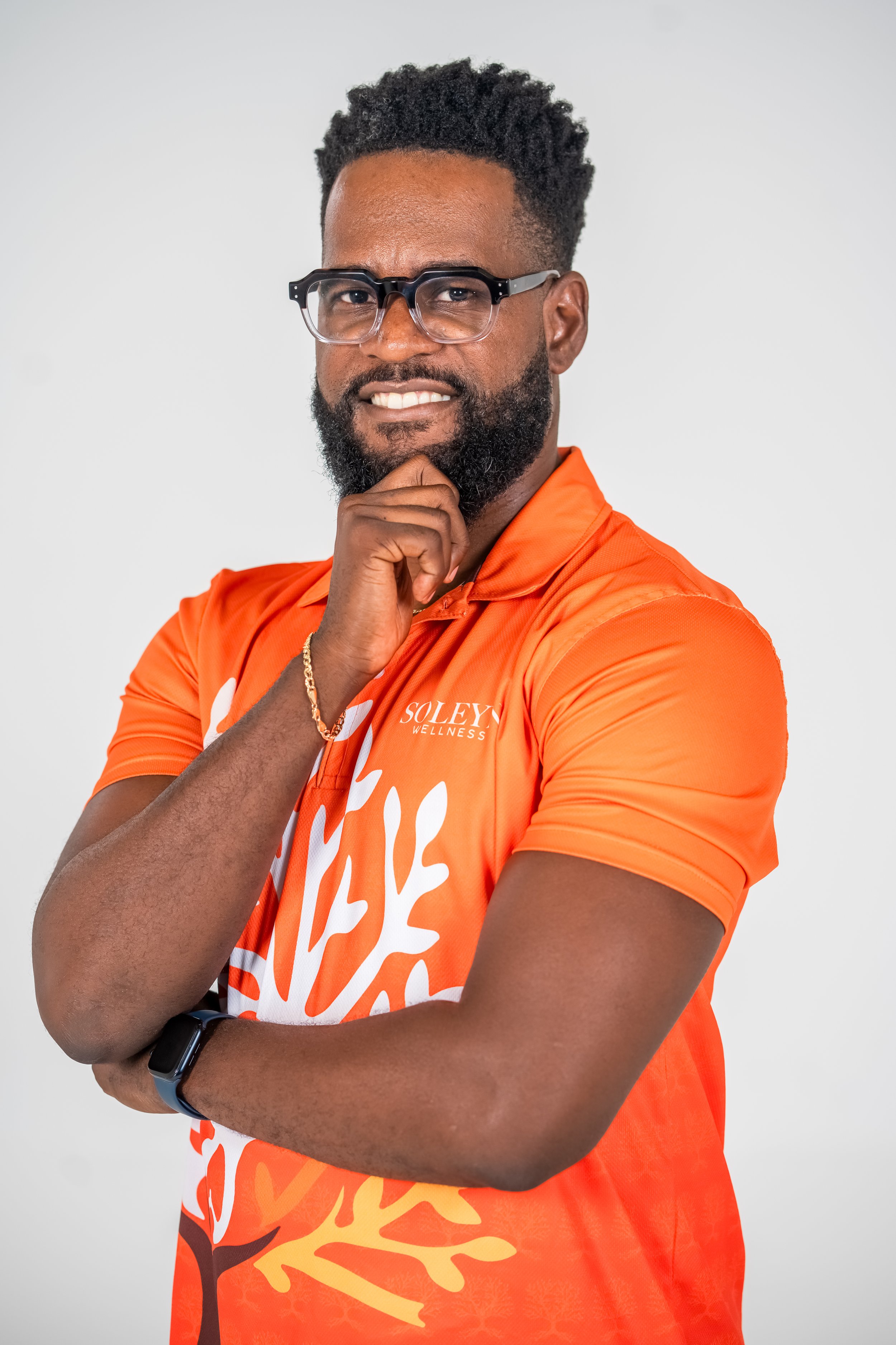 A man with a beard and glasses posing with his hand on his chin, wearing an orange shirt with white and brown design, against a plain light background.