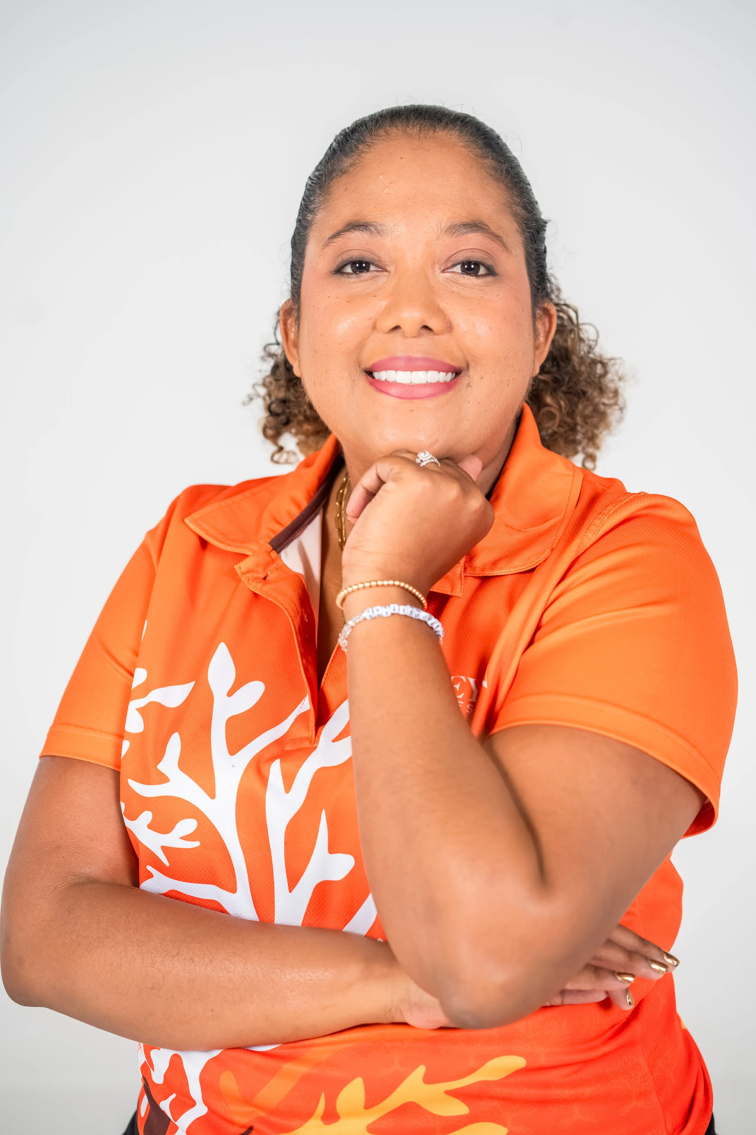 Portrait of a woman with curly hair, wearing an orange sports jersey, smiling with her hand resting on her chin, against a plain white background.