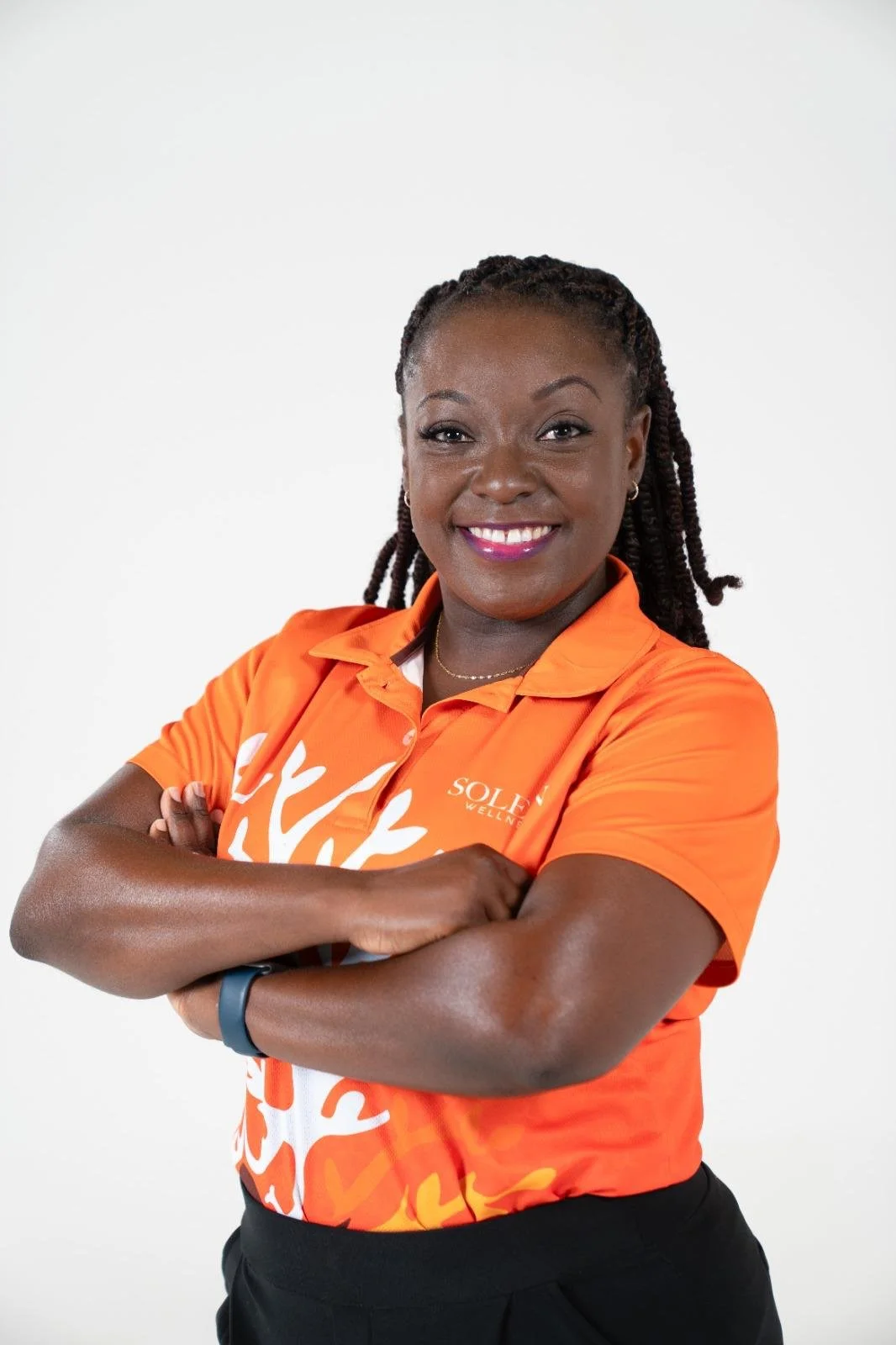 A woman smiling with arms crossed, wearing an orange polo shirt with white floral design and the logo 'Sole Wellness', against a plain white background.