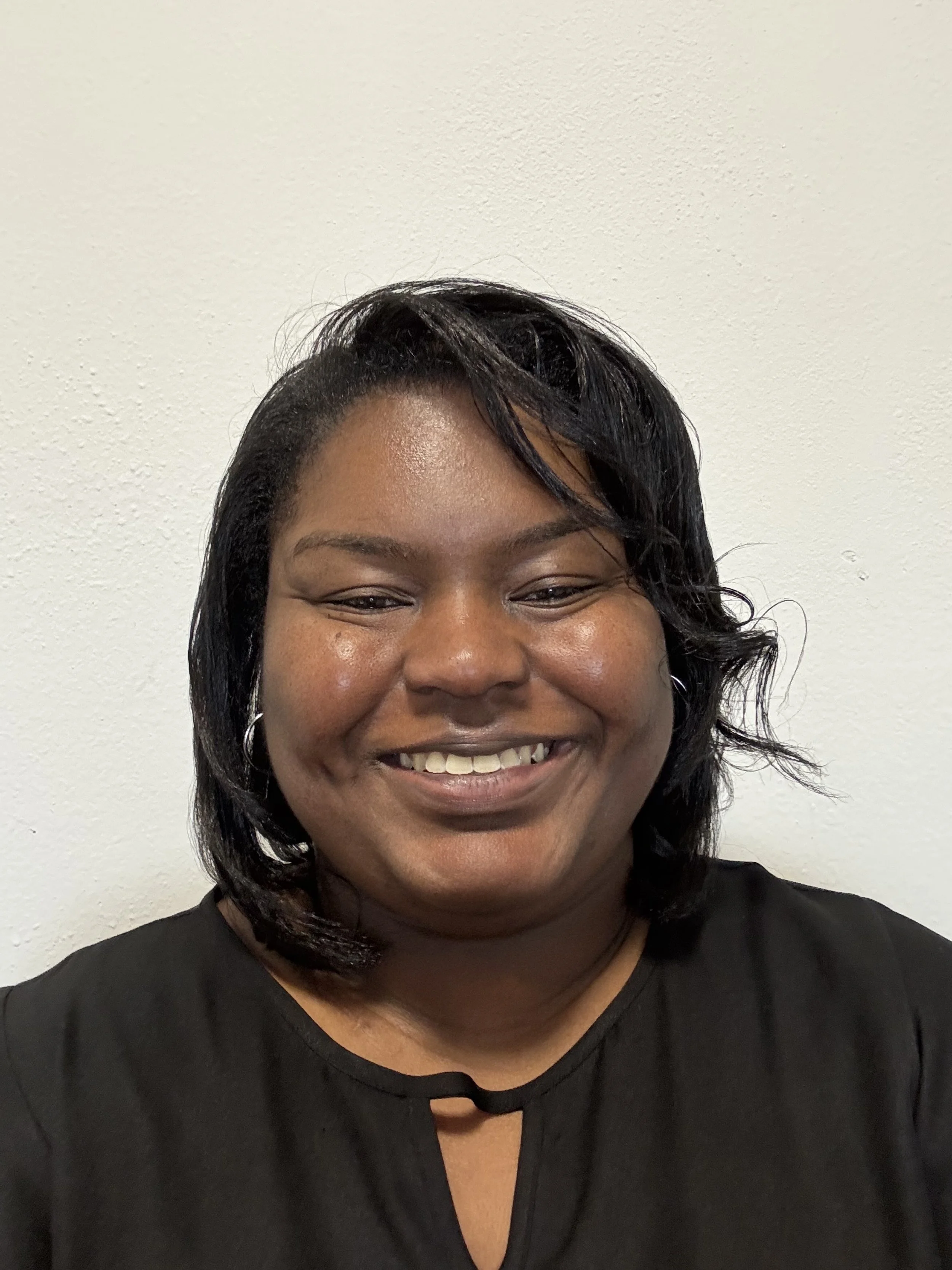 A smiling woman with shoulder-length black hair, wearing hoop earrings and a black top, standing against a plain white wall.
