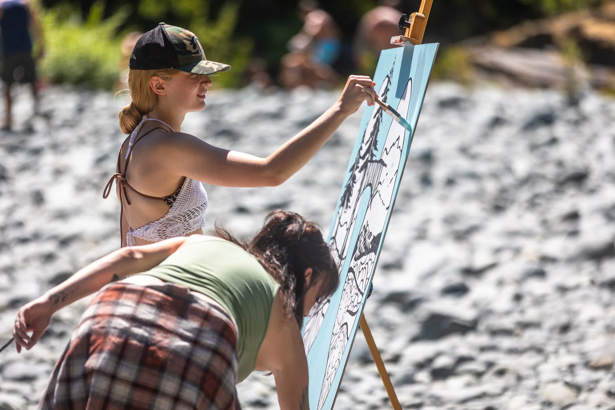 DiRT & Glitter youth intern Bailey works on a painting with artist Jessica Cherry during the 2025 Breathing Bridges community event in Del Norte county, CA on the Smith River