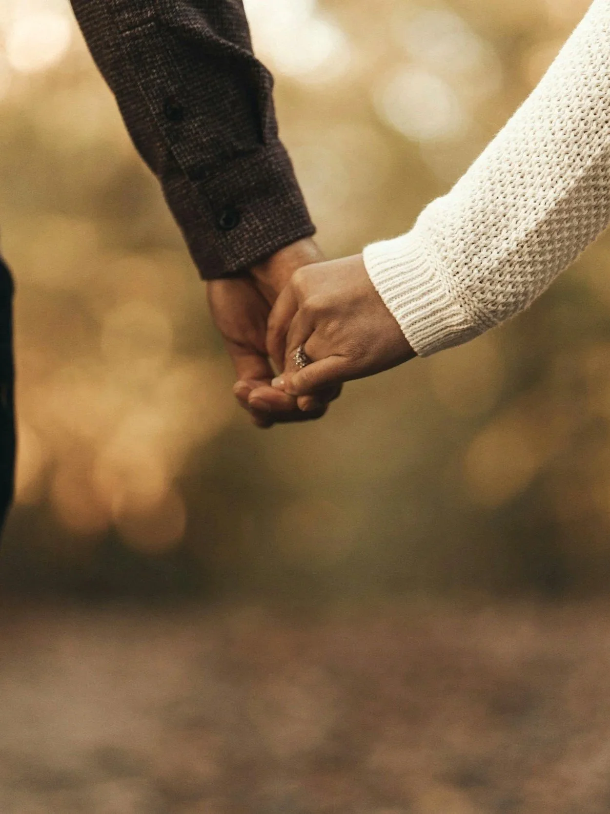 Close-up of a couple holding hands, with one wearing a dark checked jacket and the other in a white knitted sweater, outdoors with a blurred background.