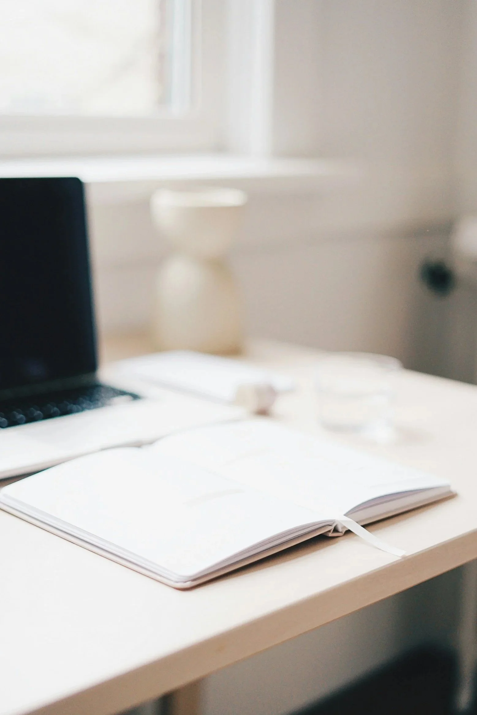 Open notebook with blank pages on a white desk, partially visible computer monitor and mouse in the background in a bright, modern workspace.