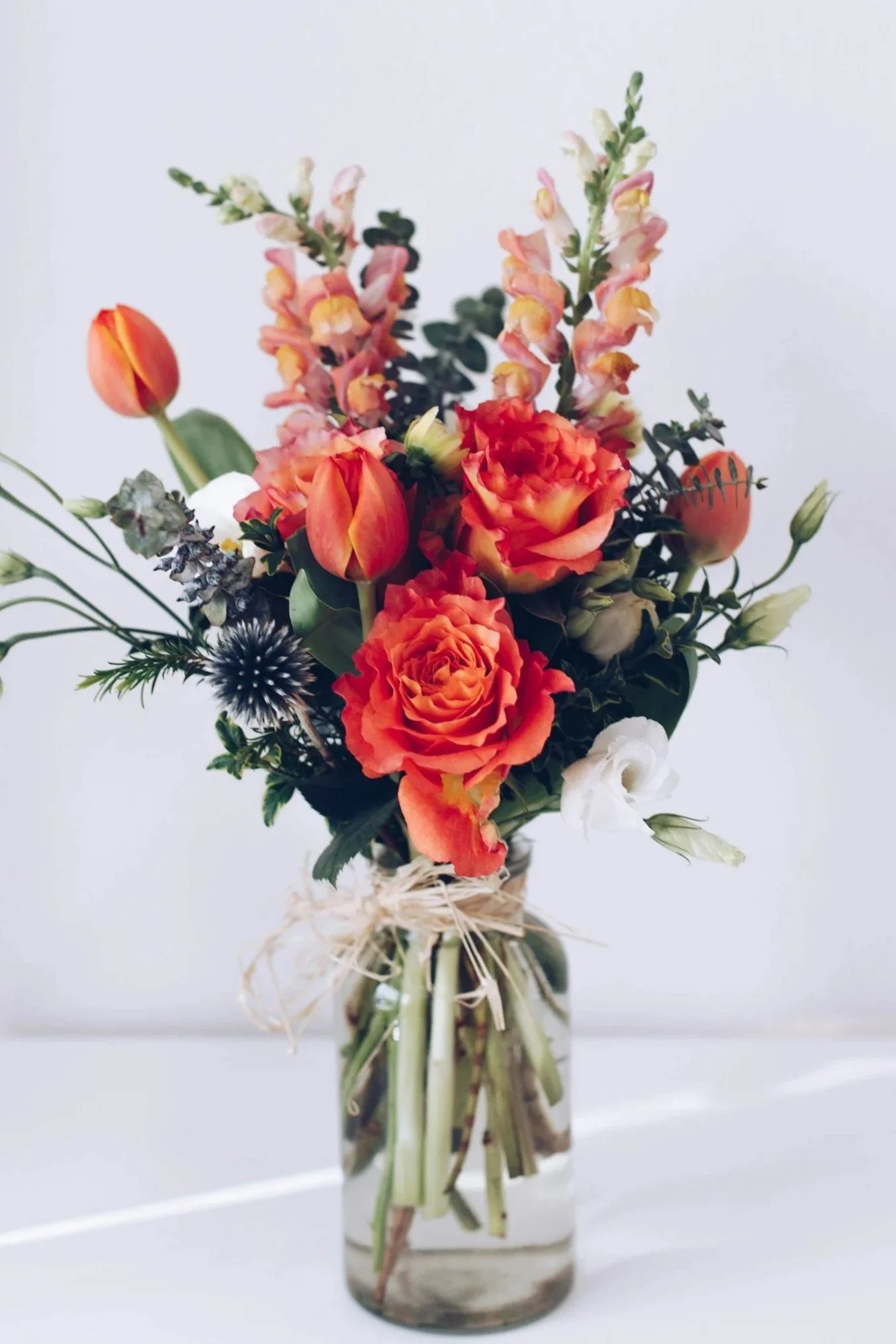 A floral arrangement with orange roses, tulips, snapdragons, and other flowers in a clear glass vase.